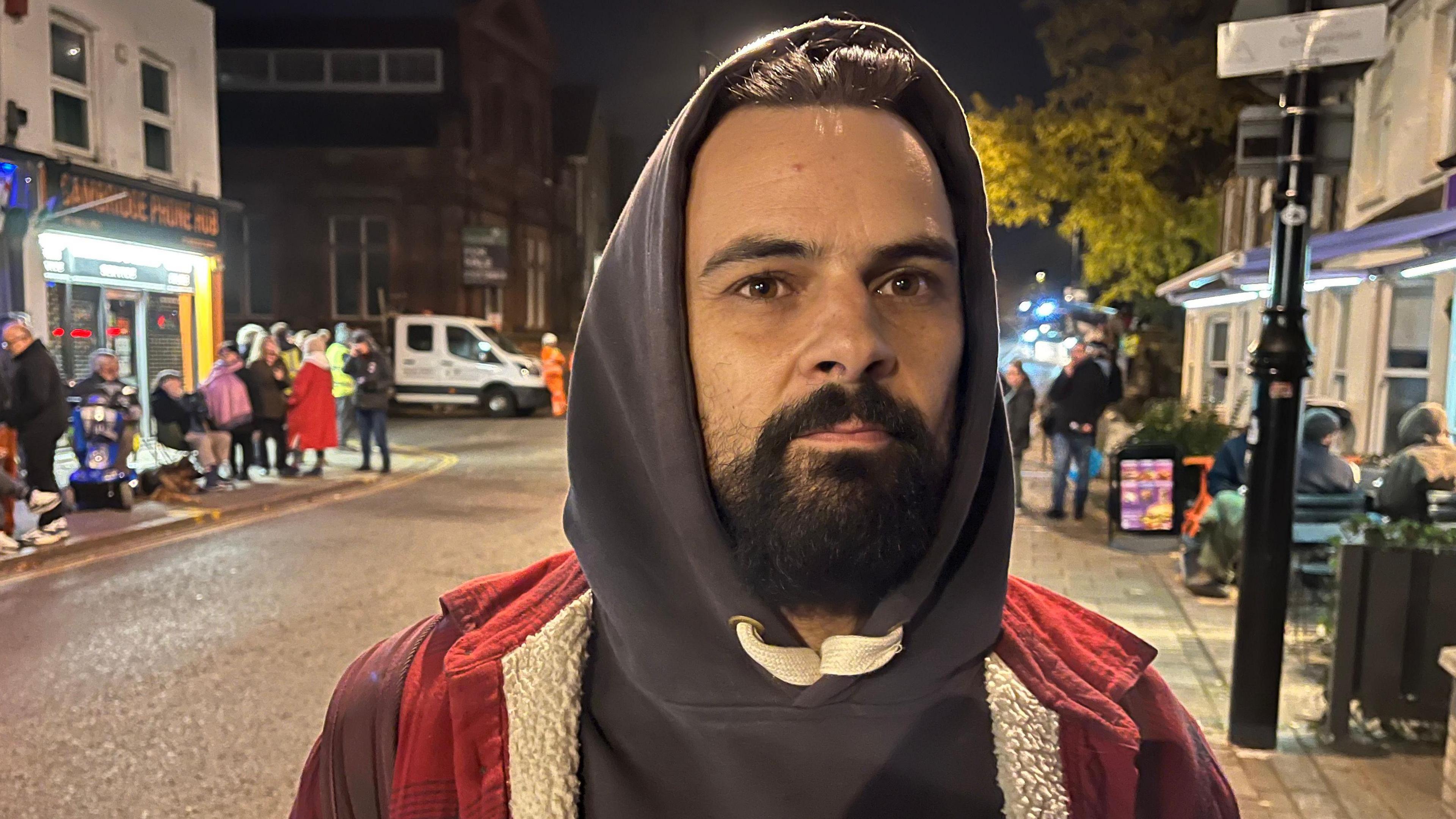 Will Bannell, has dark hair and dark facial hair. He is wearing a red jacket and has a grey hoodie in with the hood covering his head. He is looking at the camera and people can be seen lining the streets behind him. 