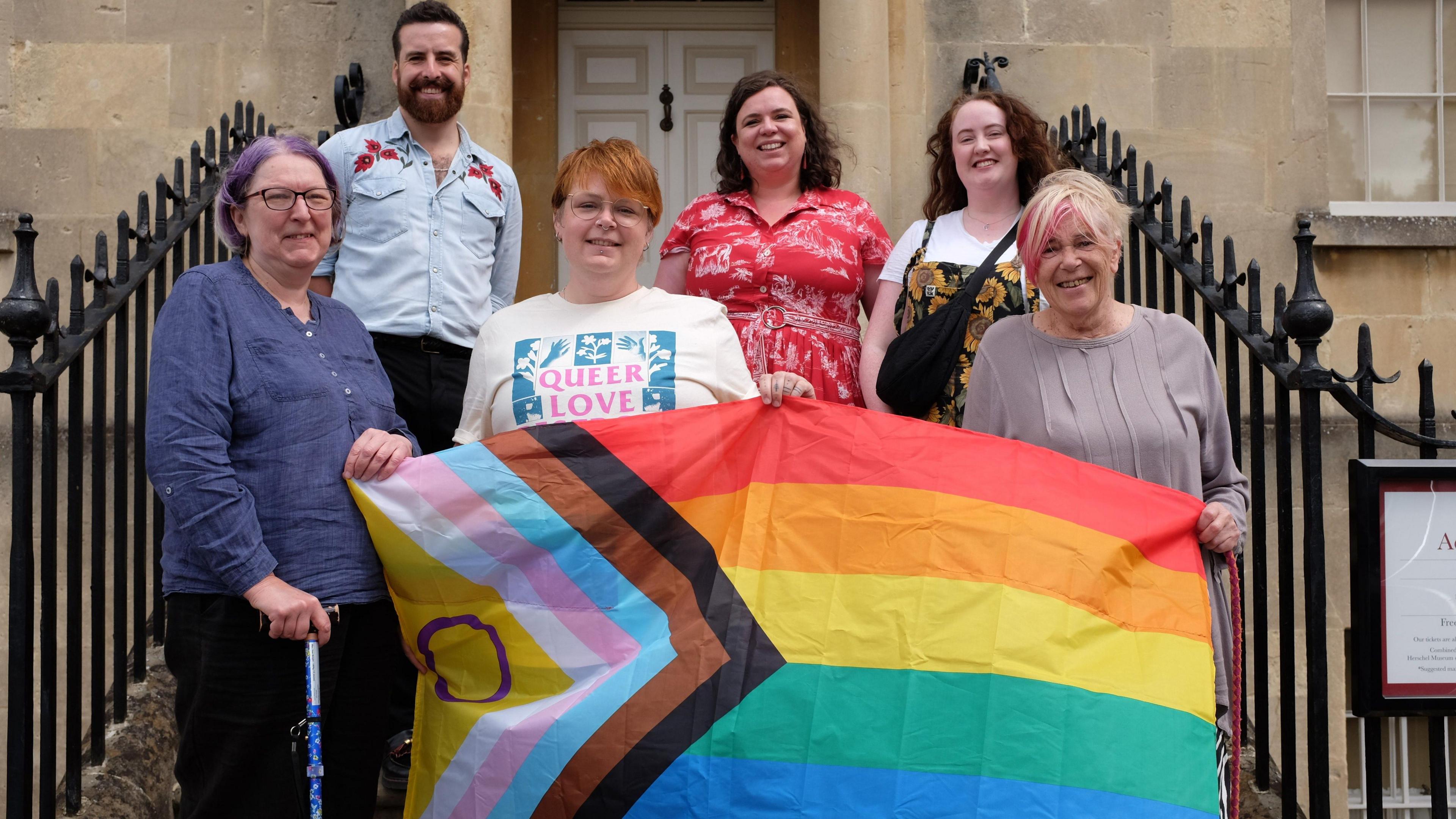 Six of the Bath Pride trustees and members of the organising committee, standing on a set of steps holding up a LGBTQ flag and smiling