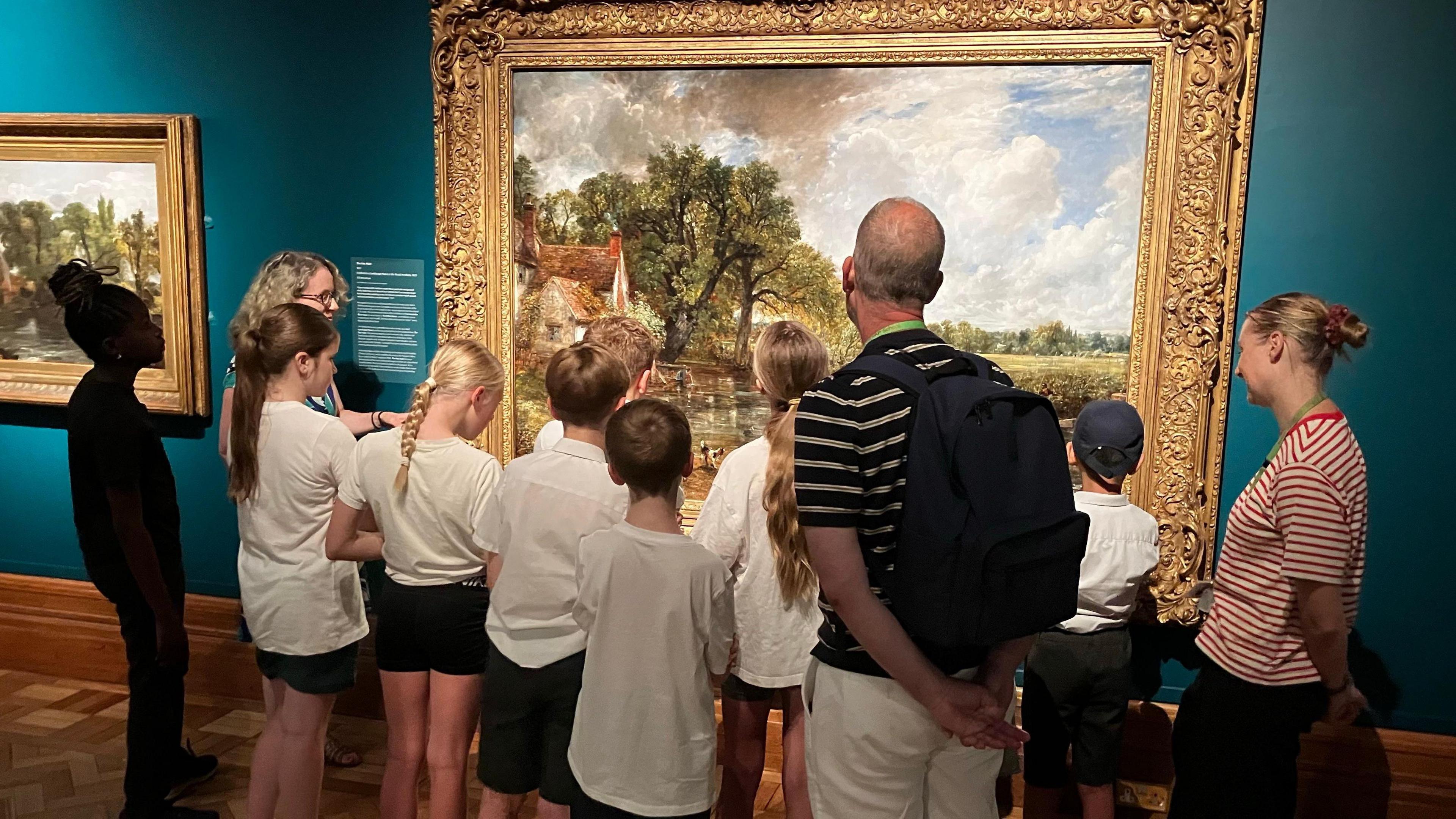 School pupils and teachers look at a large oil painting in an art exhibition. Their backs are to the camera. 