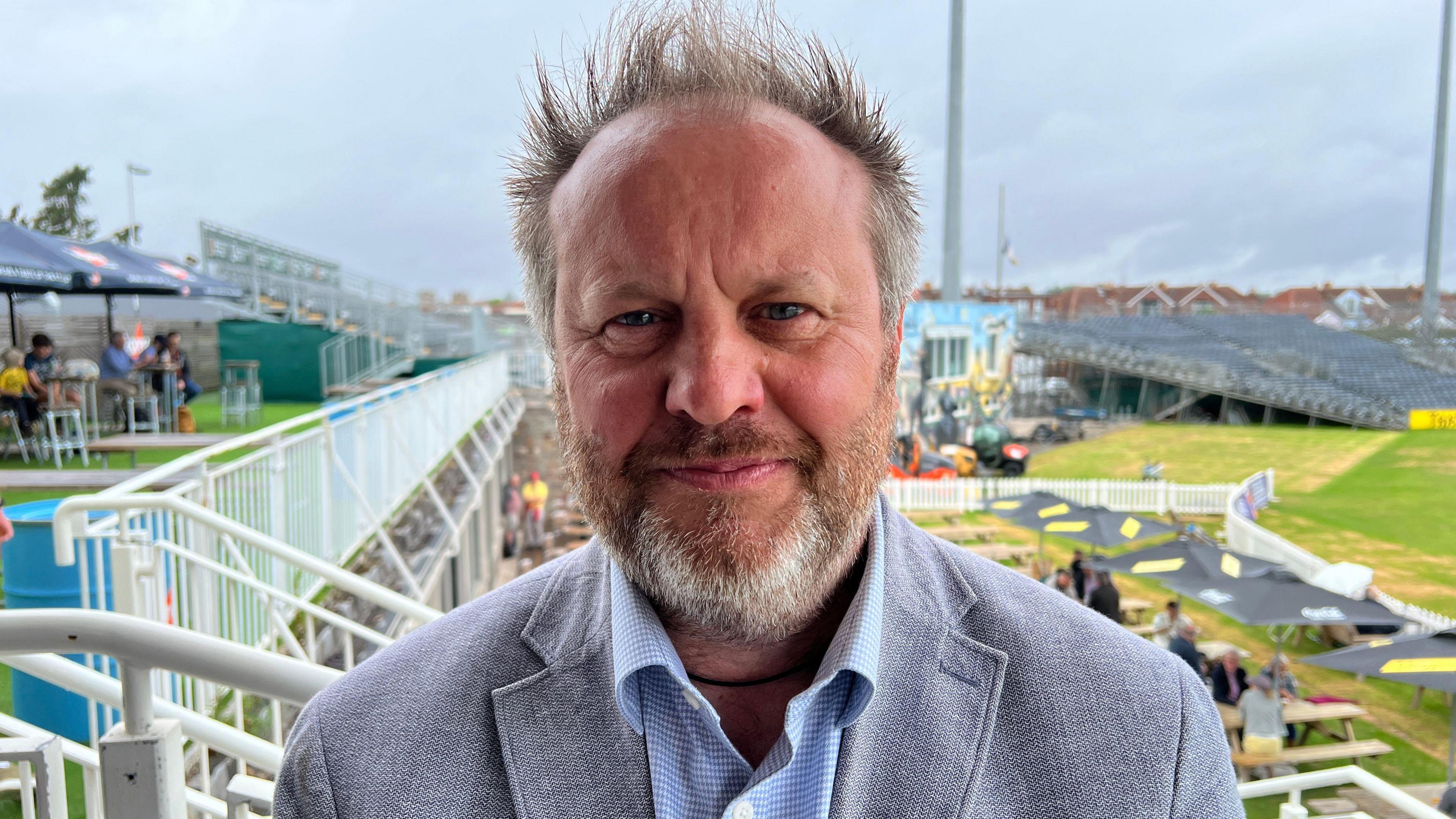 Neil Priscott. He has short grey hair and facial hair. He is wearing a blue check shirt and a grey suit jacket. He is looking at the camera and smiling.