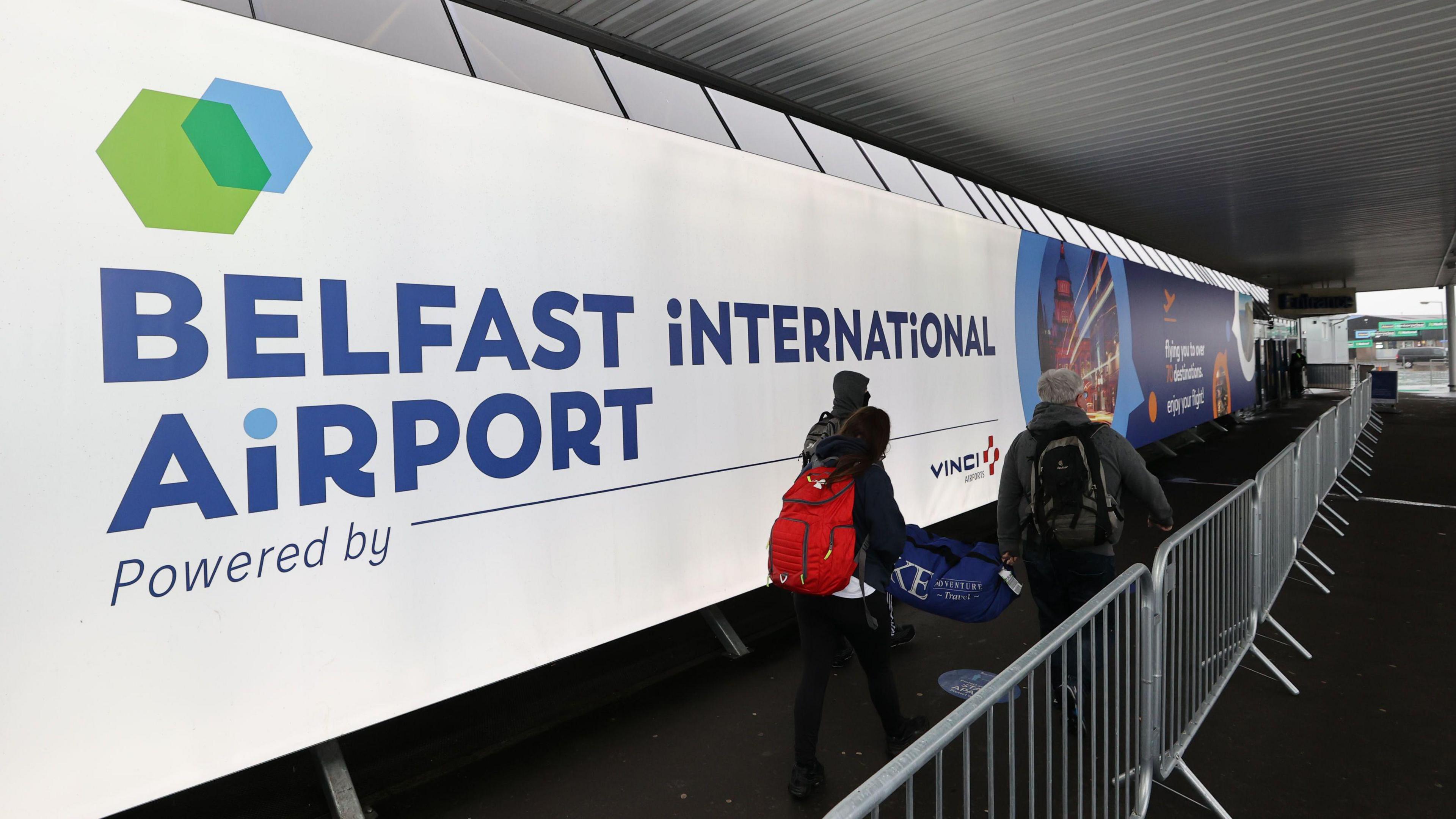 A sign reads "Belfast International Airport" - passengers walk past on their way to the terminal