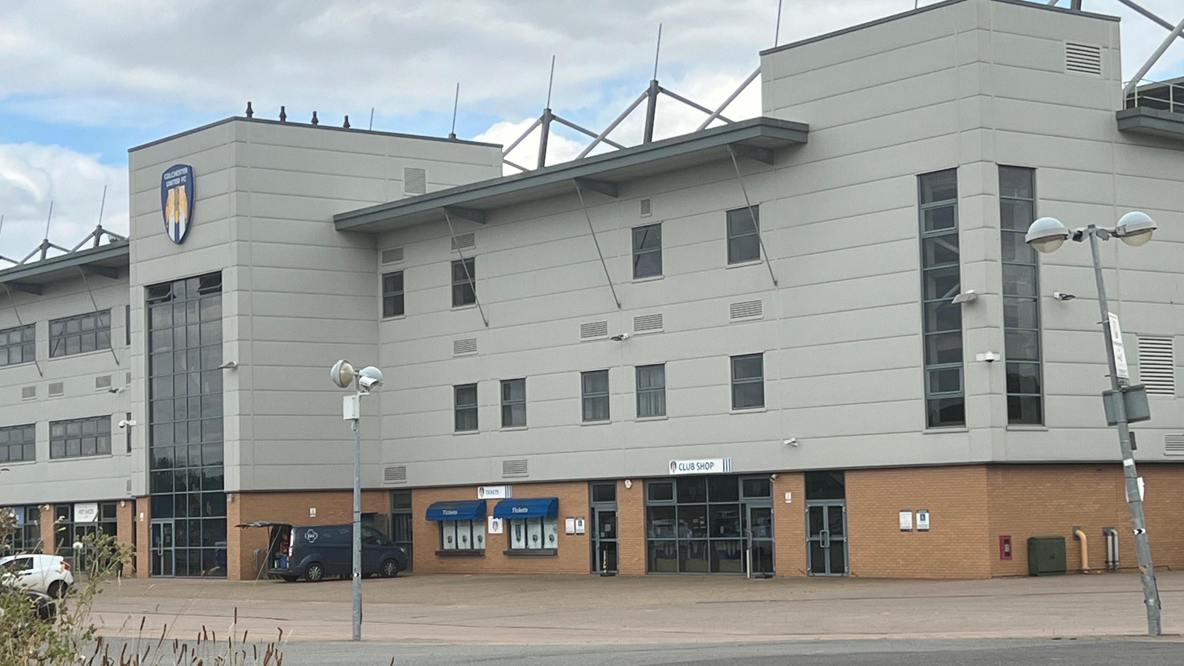 Colchester United's Community Stadium. The front of the stadium is mainly grey with some red brick at the bottom. Large windows can be seen along with several vehicles parked outside.