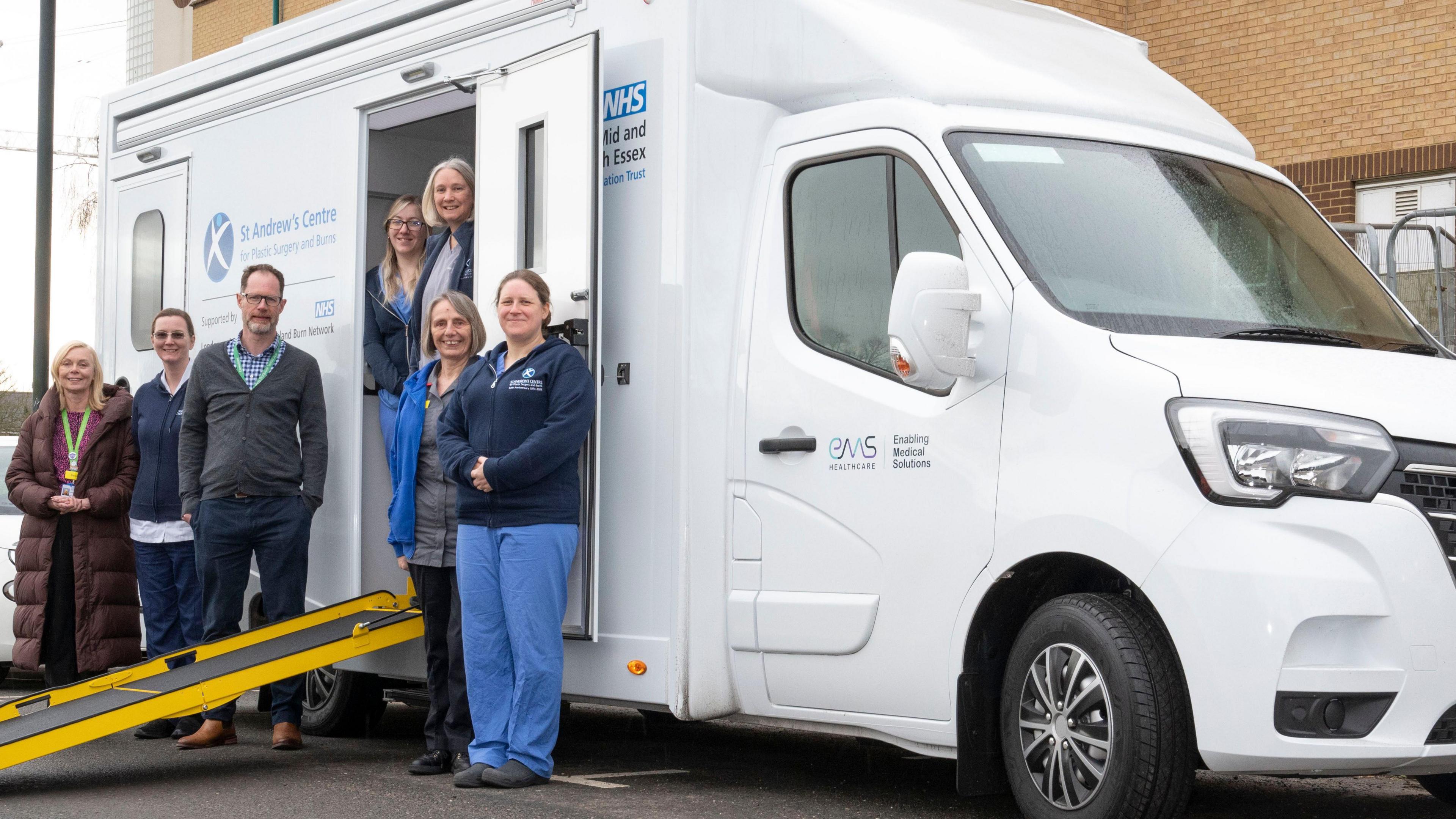 A white van is seen, with the front of the car to the right of the picture. There is a door in the middle of the van, which is open and a yellow ramp is out on the floor. 7 people are seen standing around the van. 