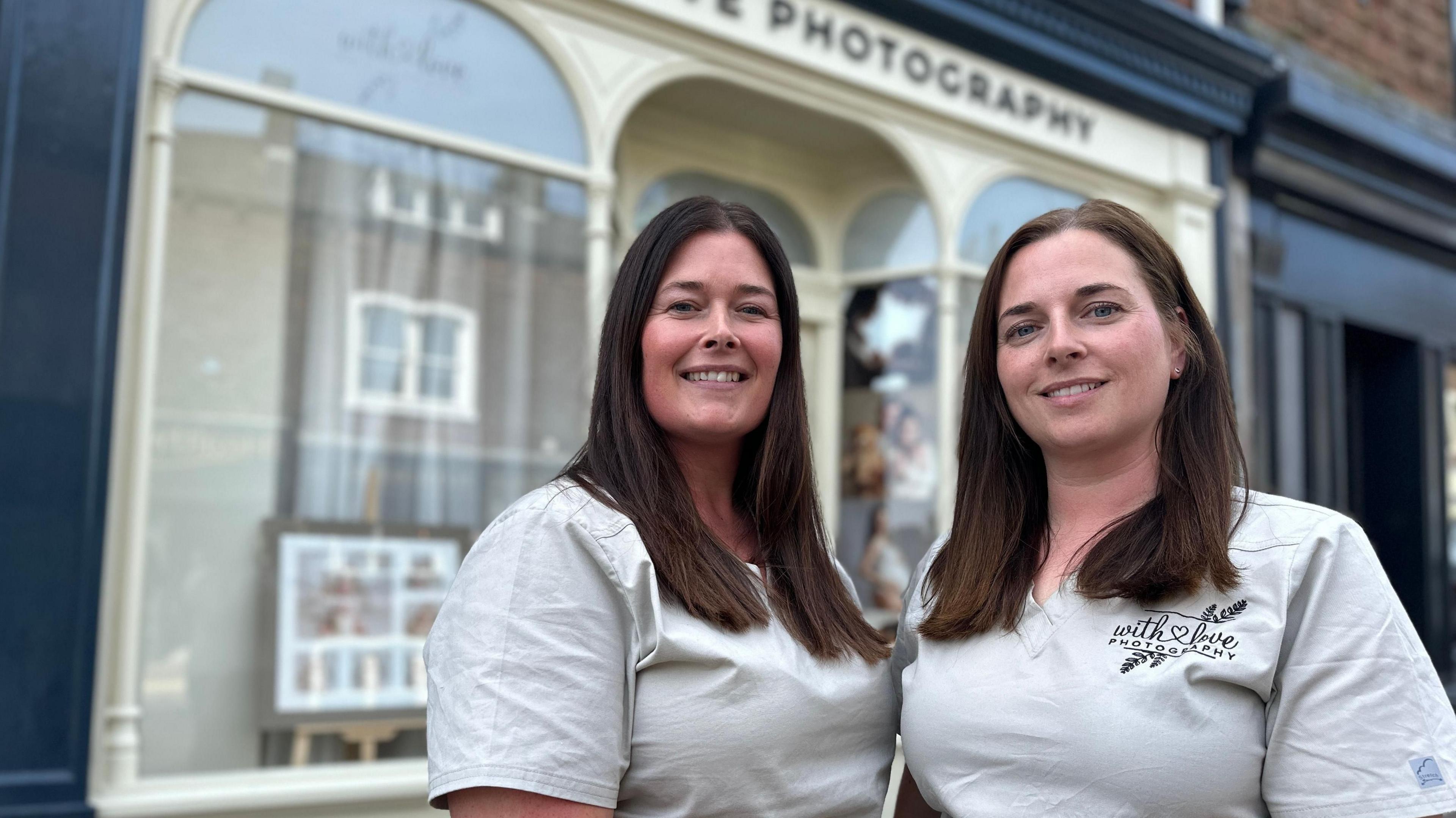 Samantha Cartwright (left) and Victoria Scott standing outside their With Love Photography studio. They both have long dark hair and are looking into the camera. Their shopfront is painted cream and has large glass windows.