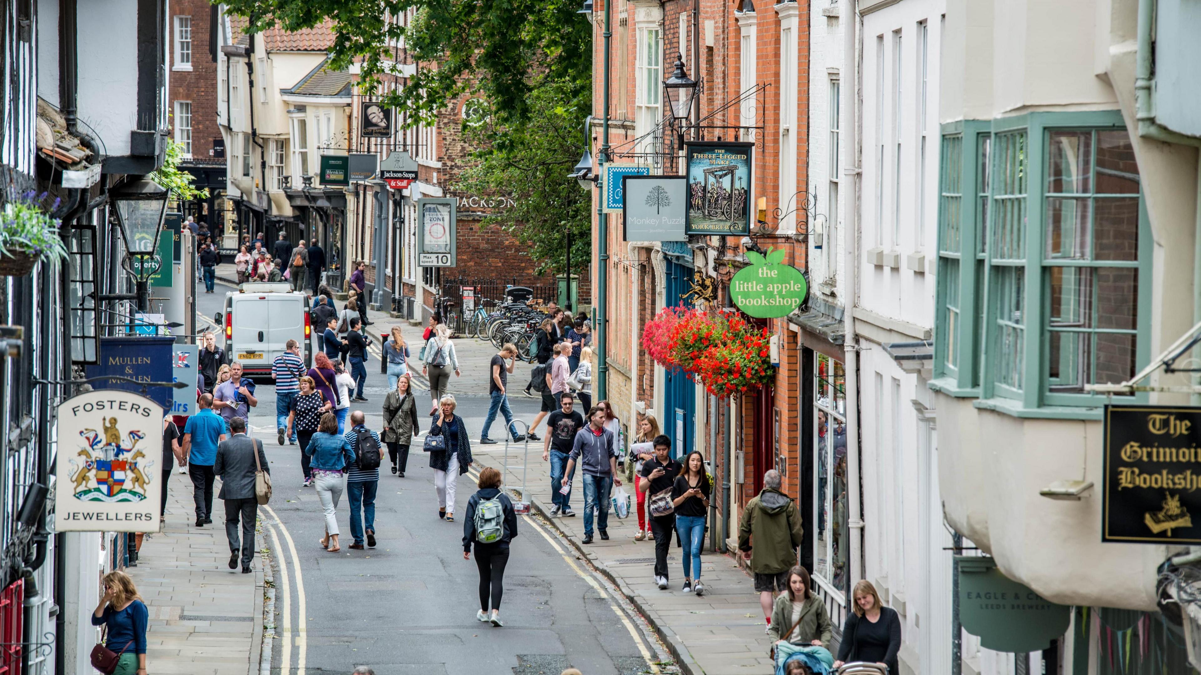 An aerial view of people walking down a narrow street in York city centre. Shop signs are seen above their respective premises.