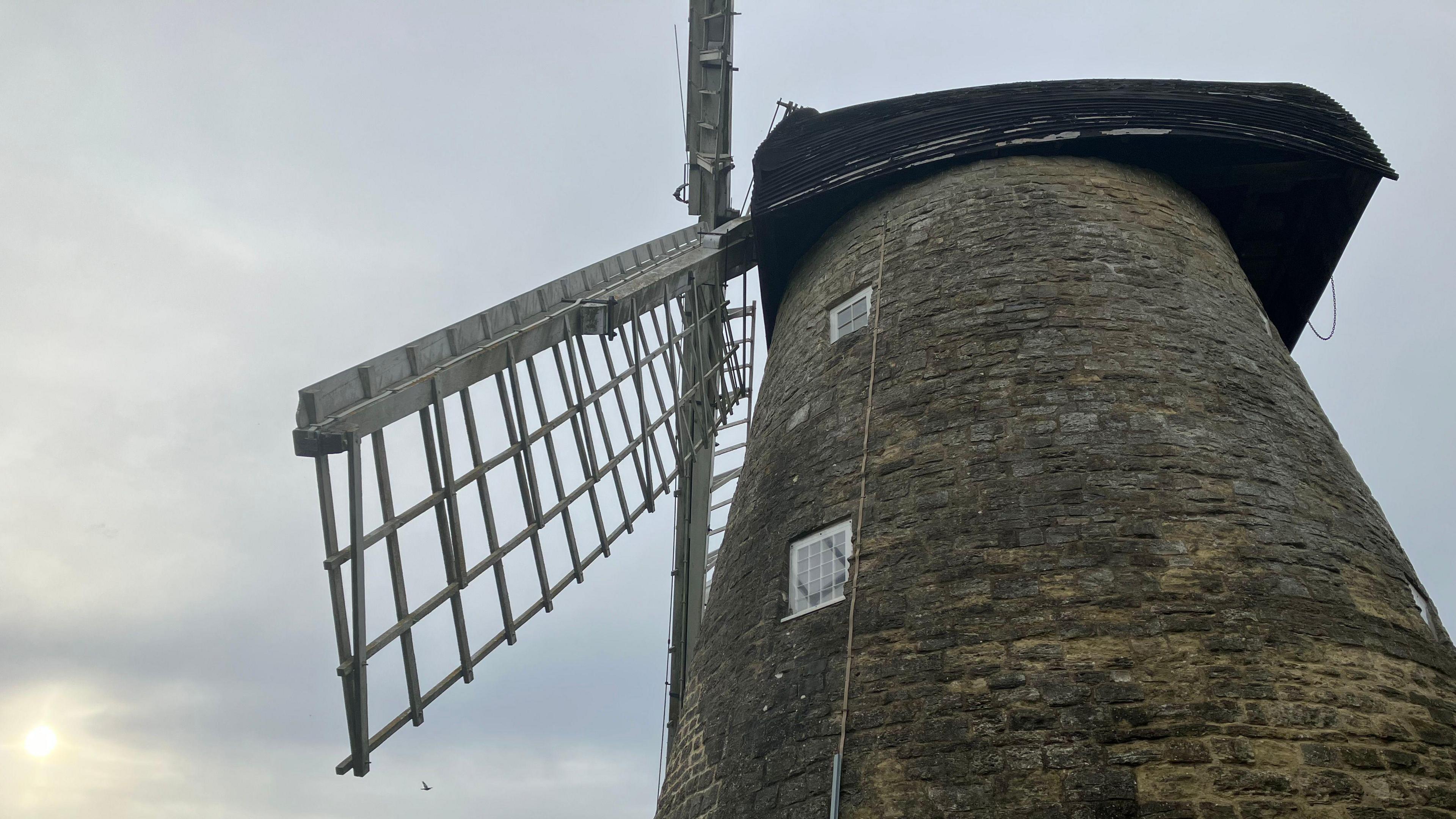 A side on picture of Bradwell windmill, where you can see part of its sails and top floors of the building. The sun is going down behind it, but the skies are grey.