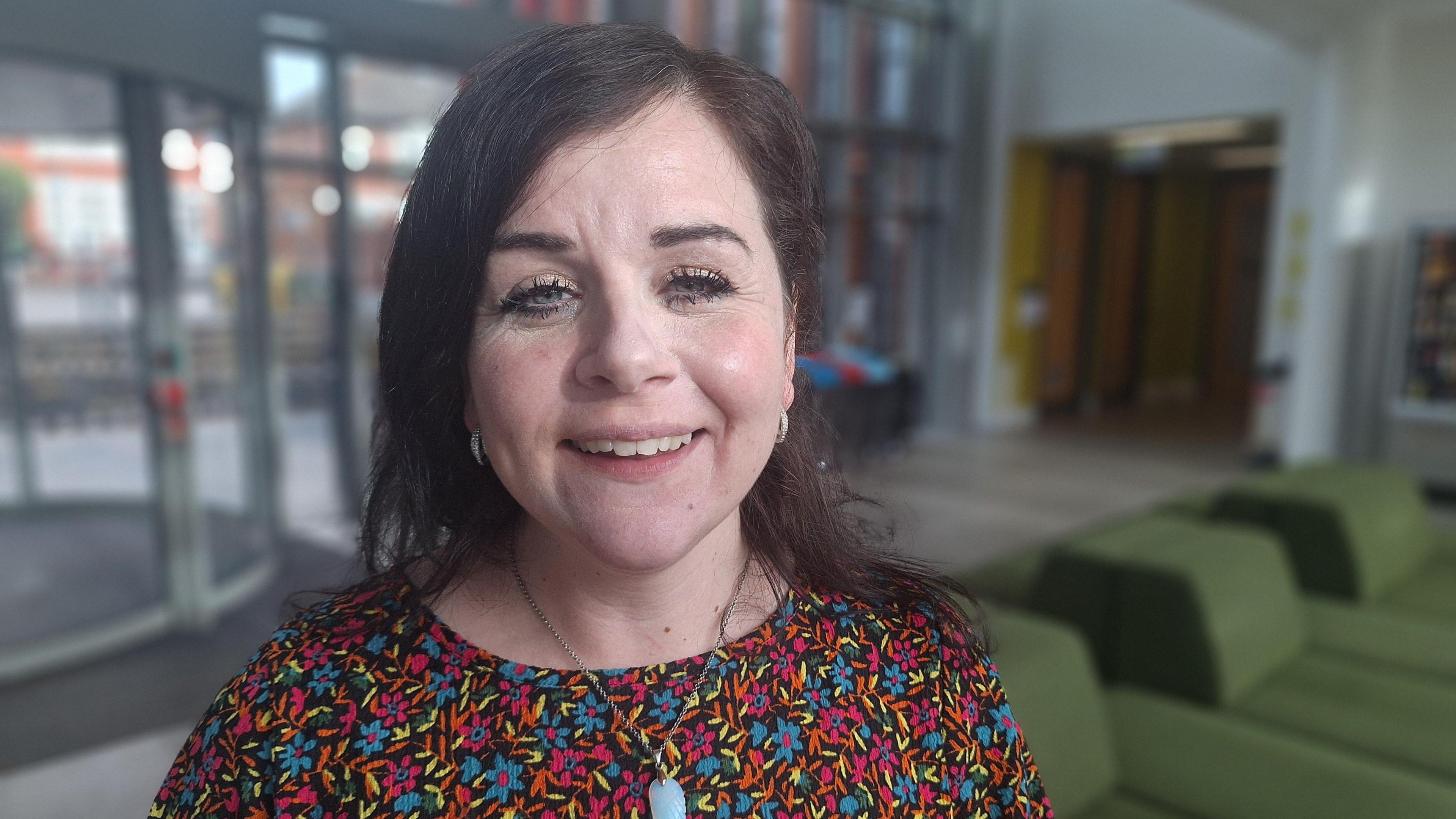Carina is looking into camera and has a big smile. She is wearing a black dress with small flowers that are blue, pink and orange. She has long black hair which is tied back and she wears a crystal pendant. She's standing in the lobby of Cardiff University's CUBRIC building where the study is taking place.