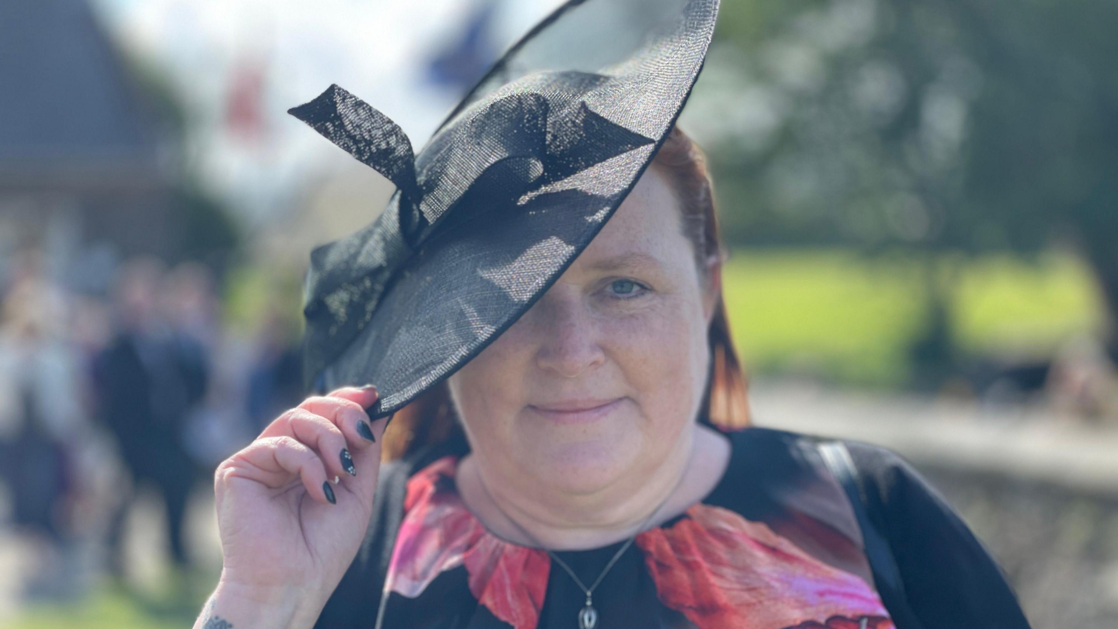 Kirstie Morphet, a woman a floral print dress, she wears a large black hat and tilts it as she smiles.
