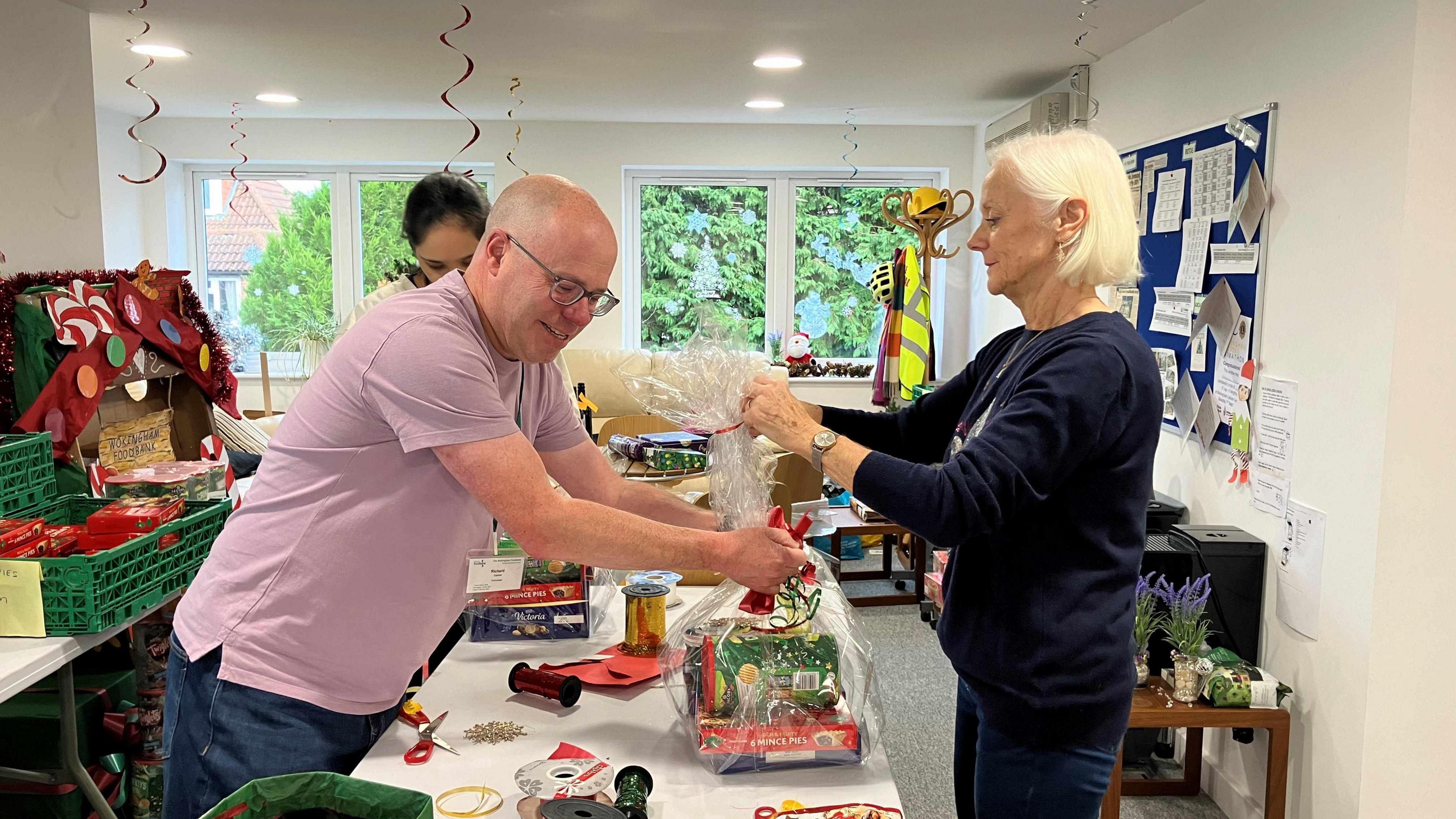 Two volunteers standing across a long table, helping each other to package up a food parcel and gift for those in need.