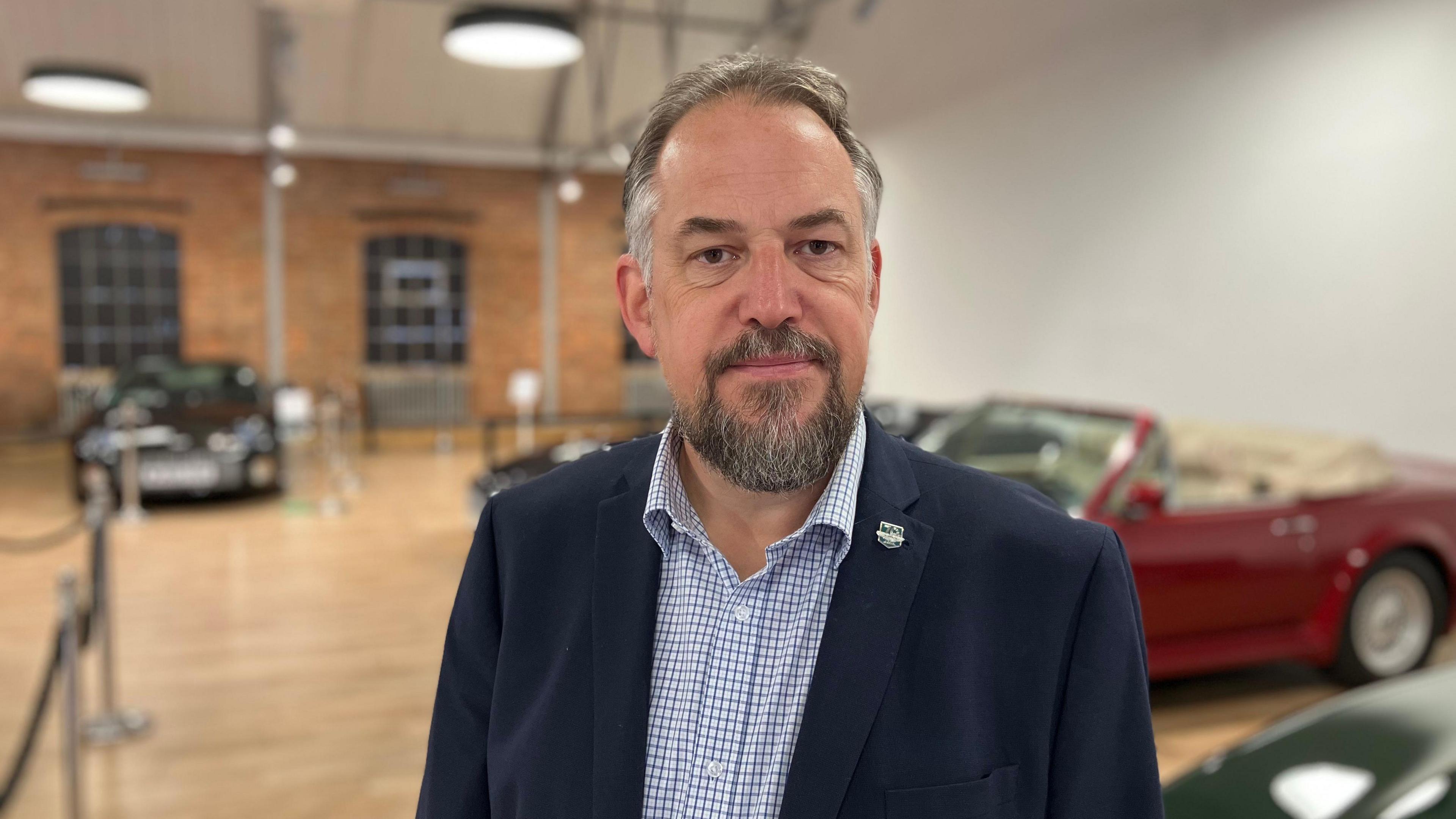 An upper body picture of Aston Martin company historian Steve Waddingham. He is looking at the camera. The background is blurred but behind him is a room with a brick wall and a plain white wall in view. There are several sports cars in the background.