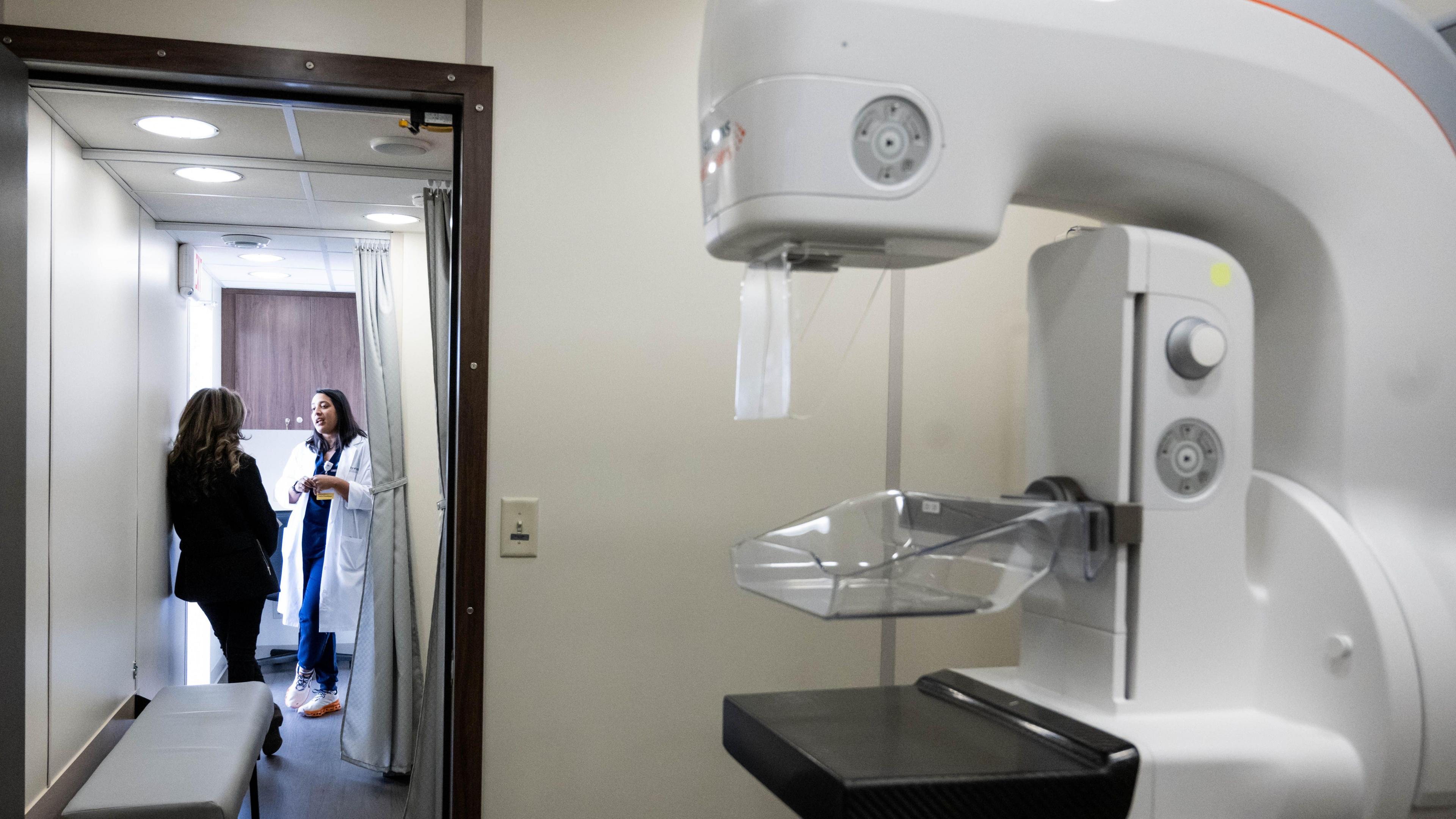 On the right is a white mammogram machine standing in a white room. On the left two women are standing talking in a corridor.