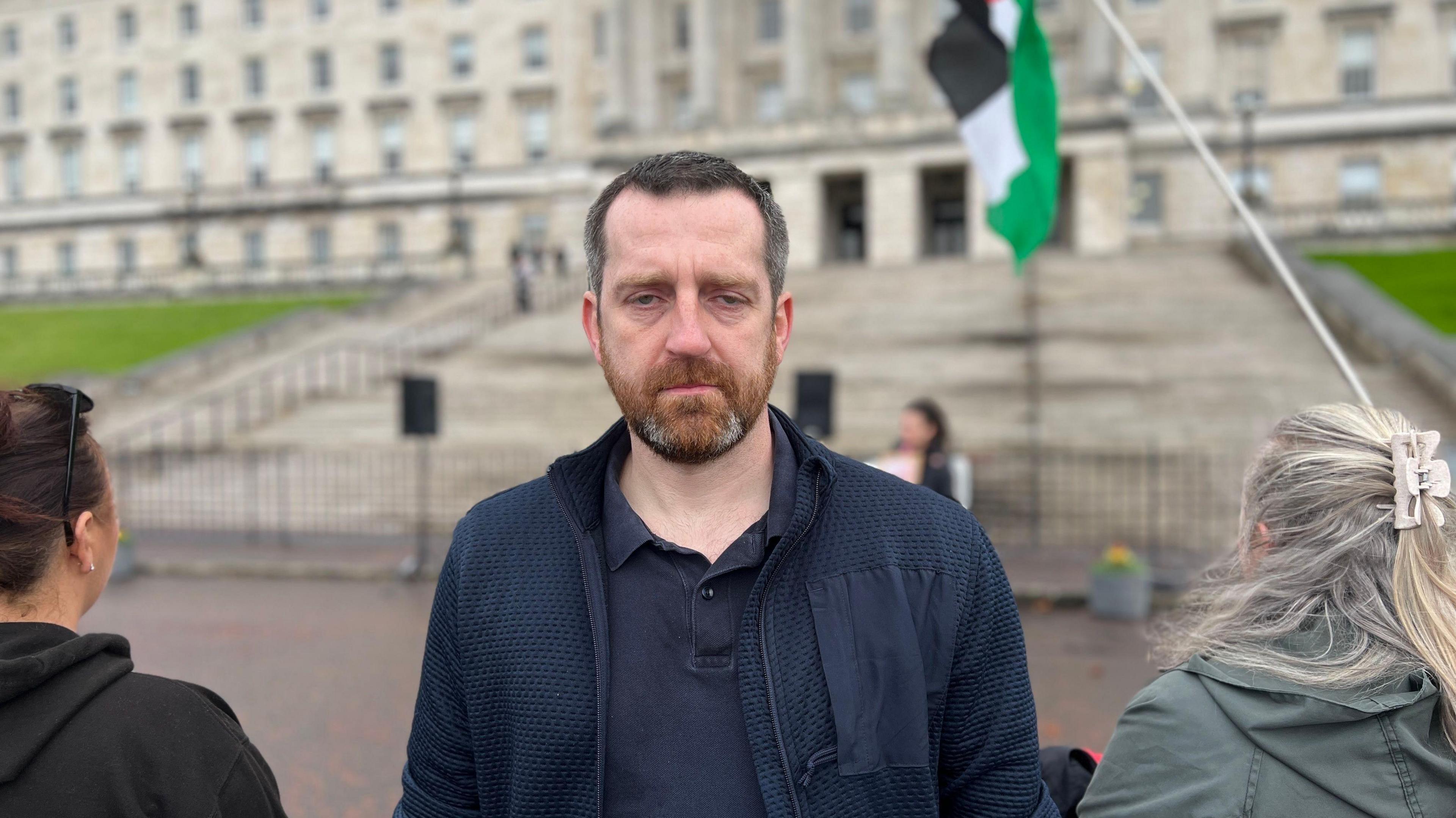A bearded man wearing a black jacket stands outside an out of focus parliament buildings. A Palestinian flag can be seen in the corner of the picture. 