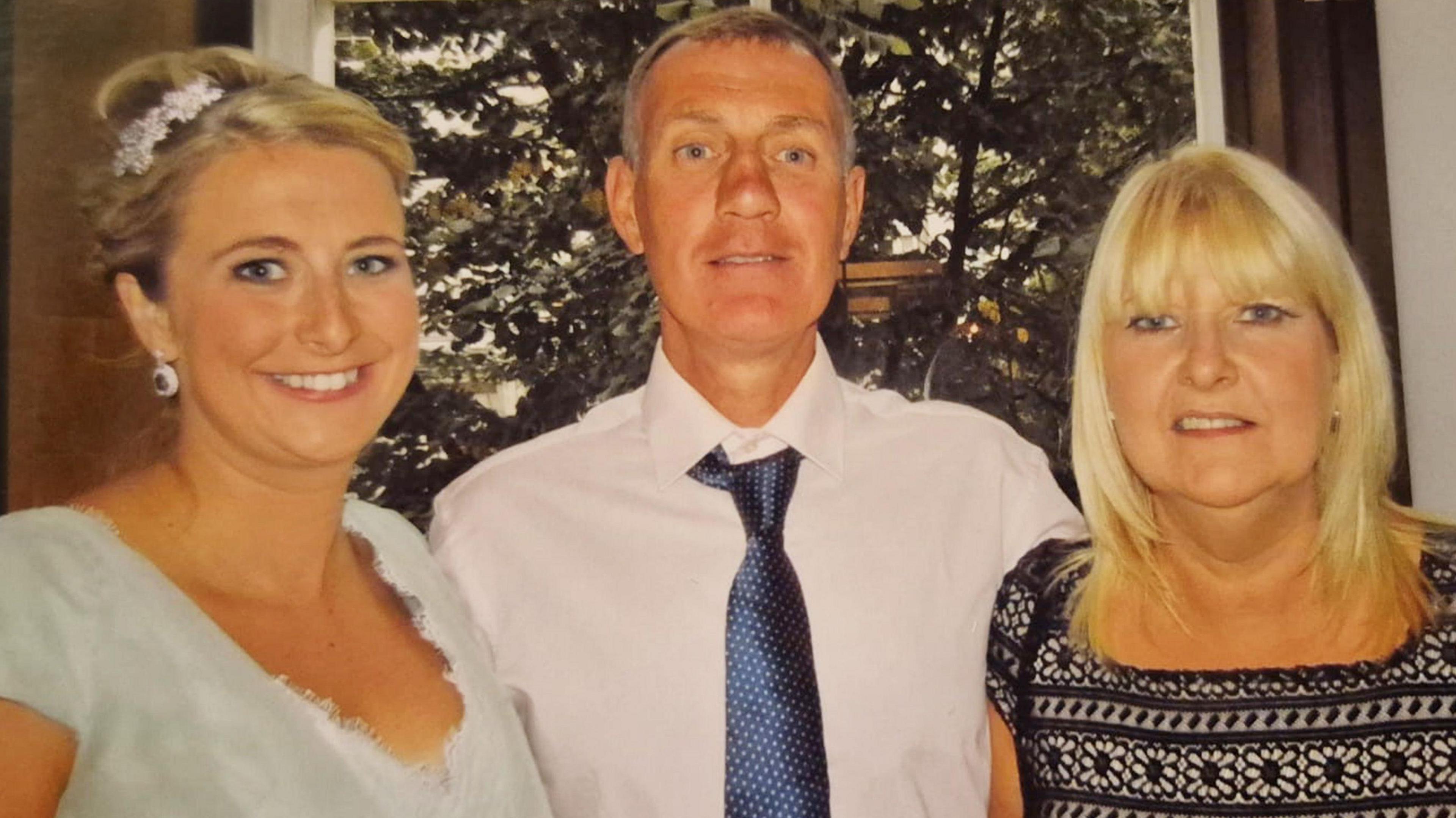 Three people stand smiling for the camera in front of a window, which shows large trees outside. To the left is a young woman in a green dress. Her blonde hair is tied up in a bun with a floral display. She has white earrings dangling. A man in a white shirt and blue tie is in the centre with an arm around the two women. He has dark hair, which is turning grey. To the right is an older woman with blonde hair which hangs by her shoulders. She is wearing a black pattered top, which appears to have flowers embroidered into the pattern.