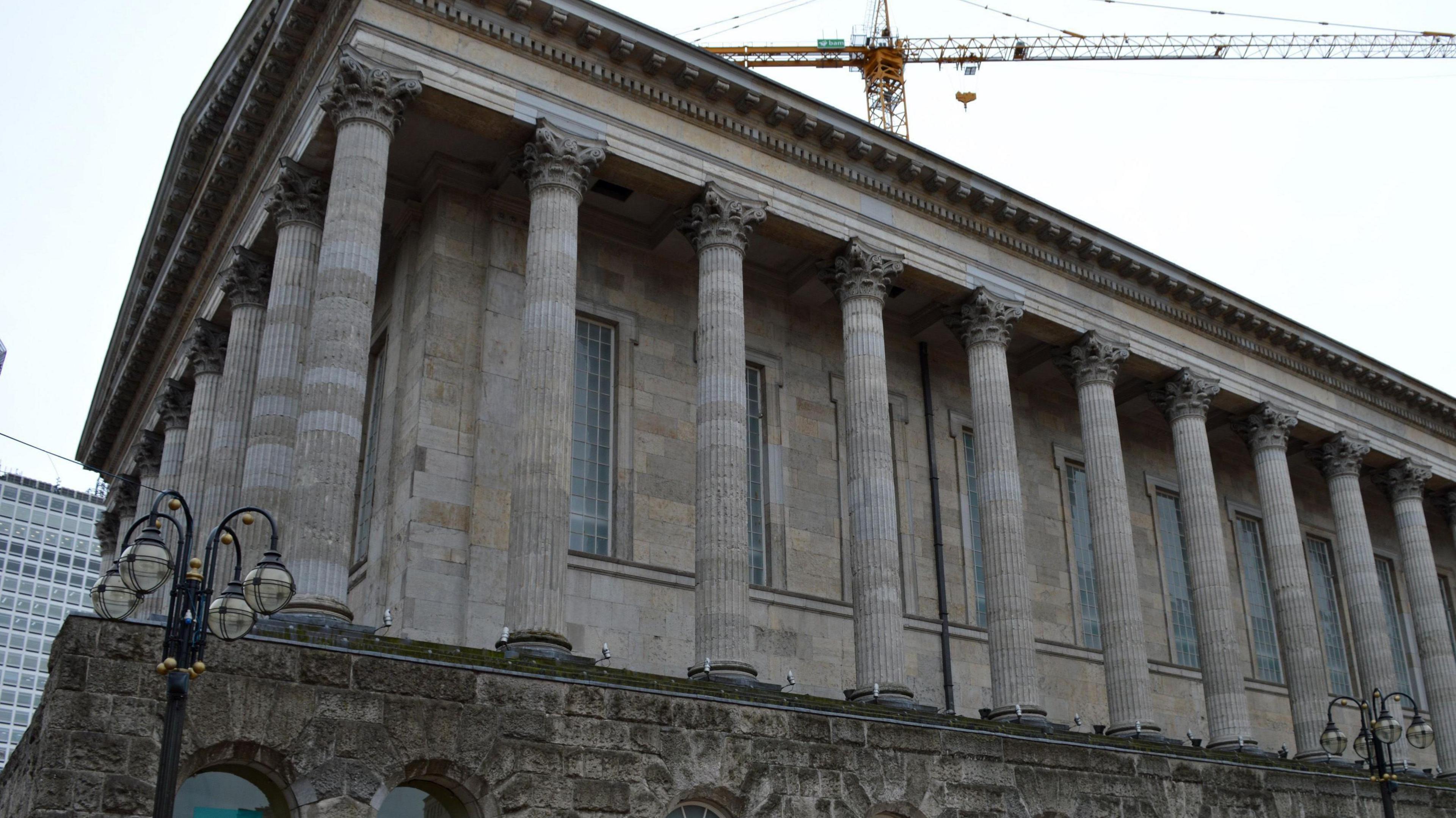 Birmingham town hall, a brick building with huge stone pillars around it. There is a brick wall with archways beneath it. A yellow crane is behind it, looming above