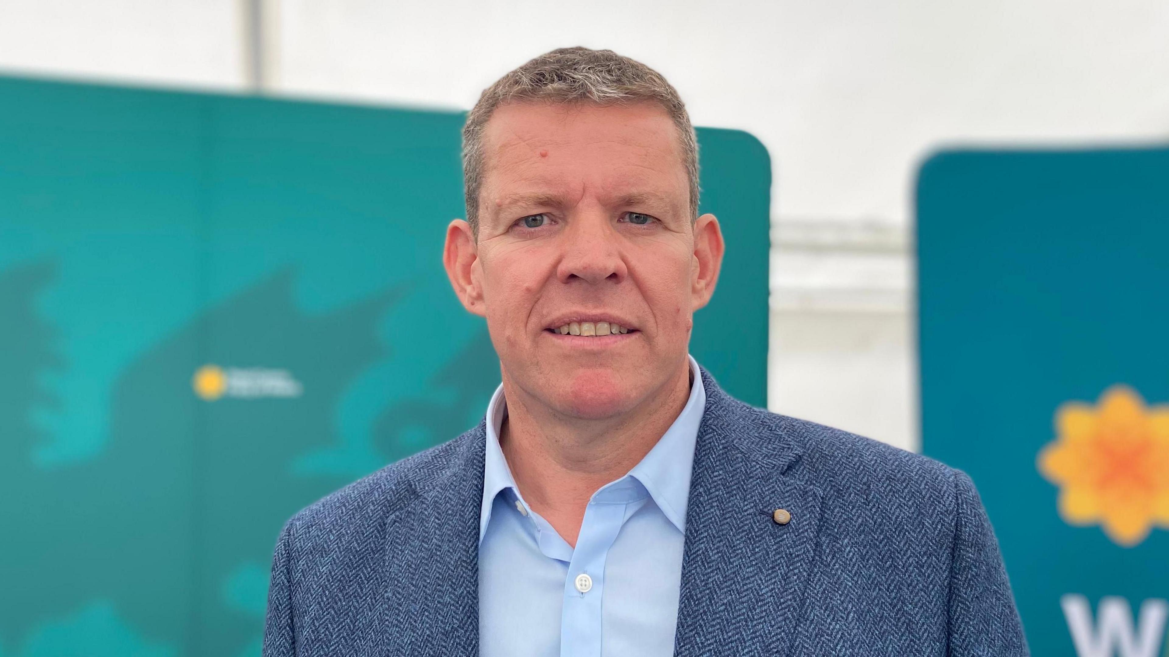 Rhub ap Iorwerth yn gwisgo crys las a siaced las. Mae'n sefyll o flaen arwyddion mawr Plaid Cymru.