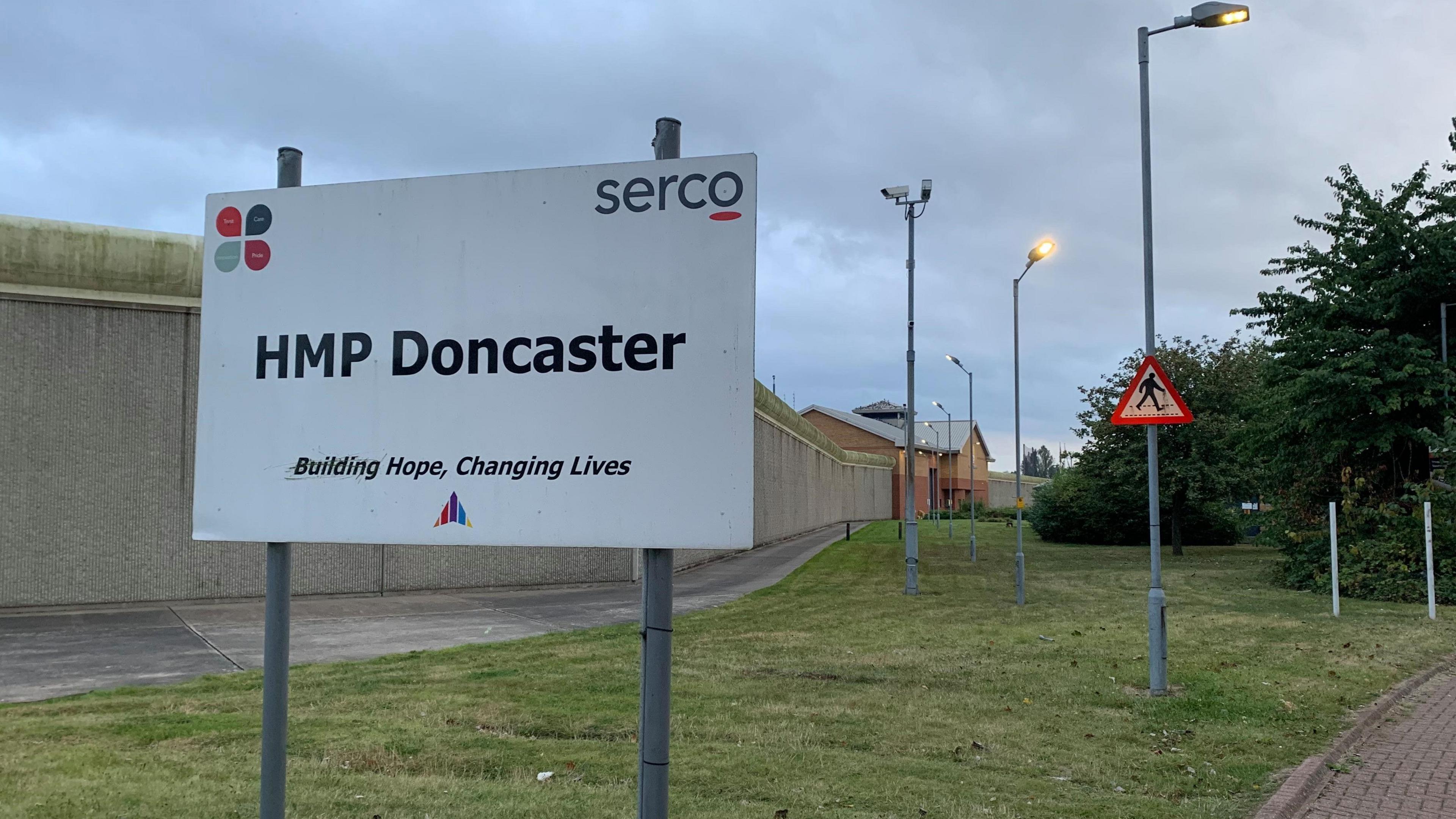 A sign reading "HMP Doncaster". In the background is a large concrete wall, surrounding the outdoor area within the prison.