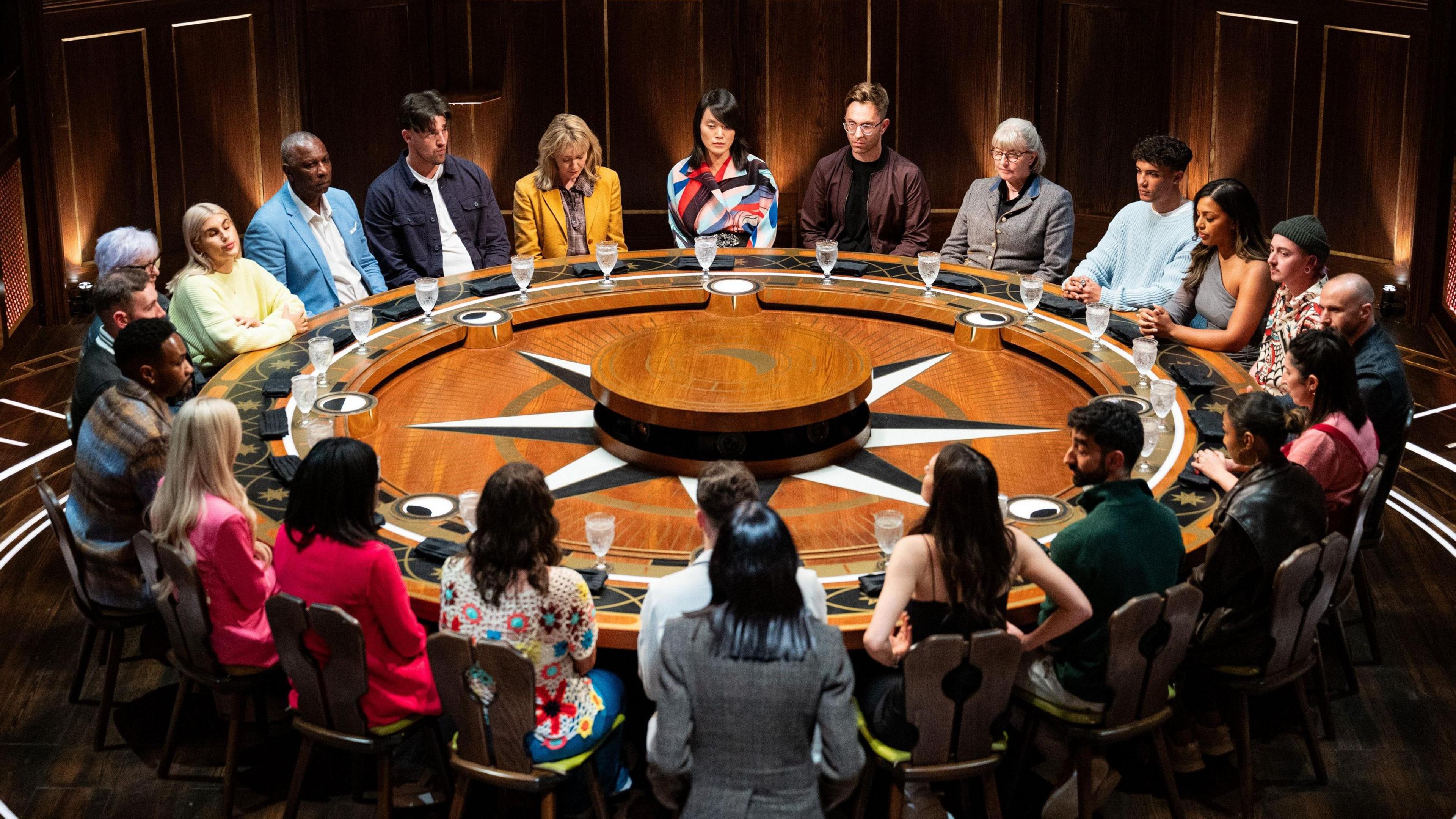 The contestants of The Traitors are sitting around a large round wooden table.