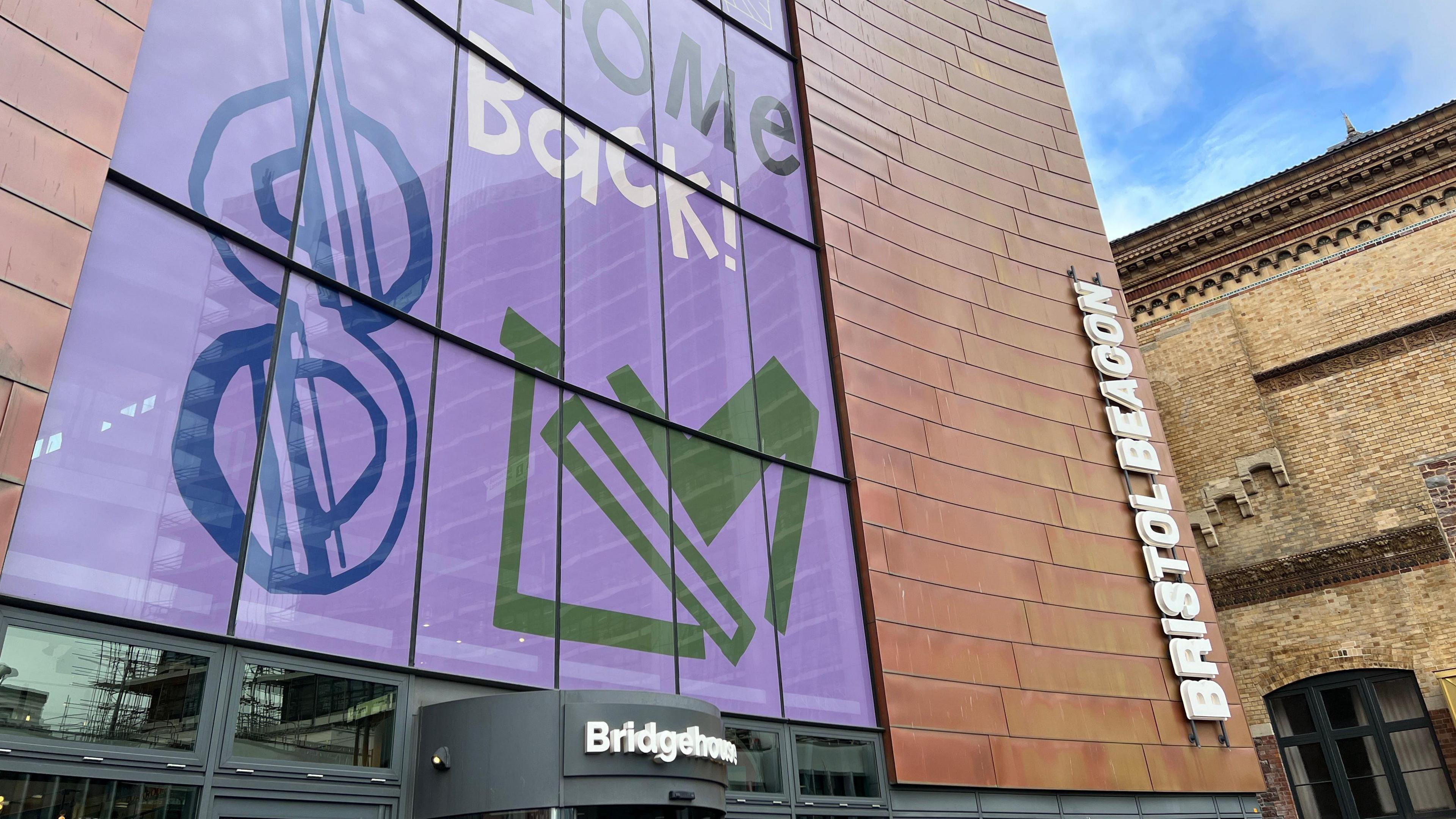 Image of Bristol Beacon. It is a red brick building with a purple front, which reads Welcome Back! 