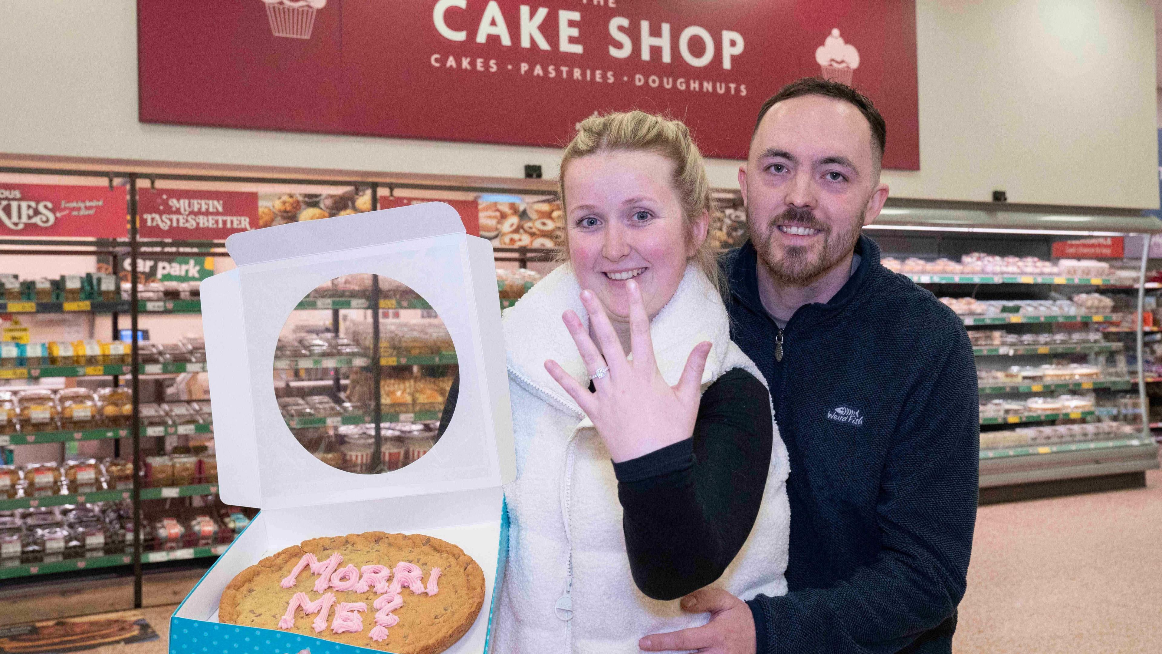 Charlie Bullock and Hannah McNaughten together at the bakery counter. Hannah is holding a giant cookie saying 'Morri me' and showing off a ring on her finger.