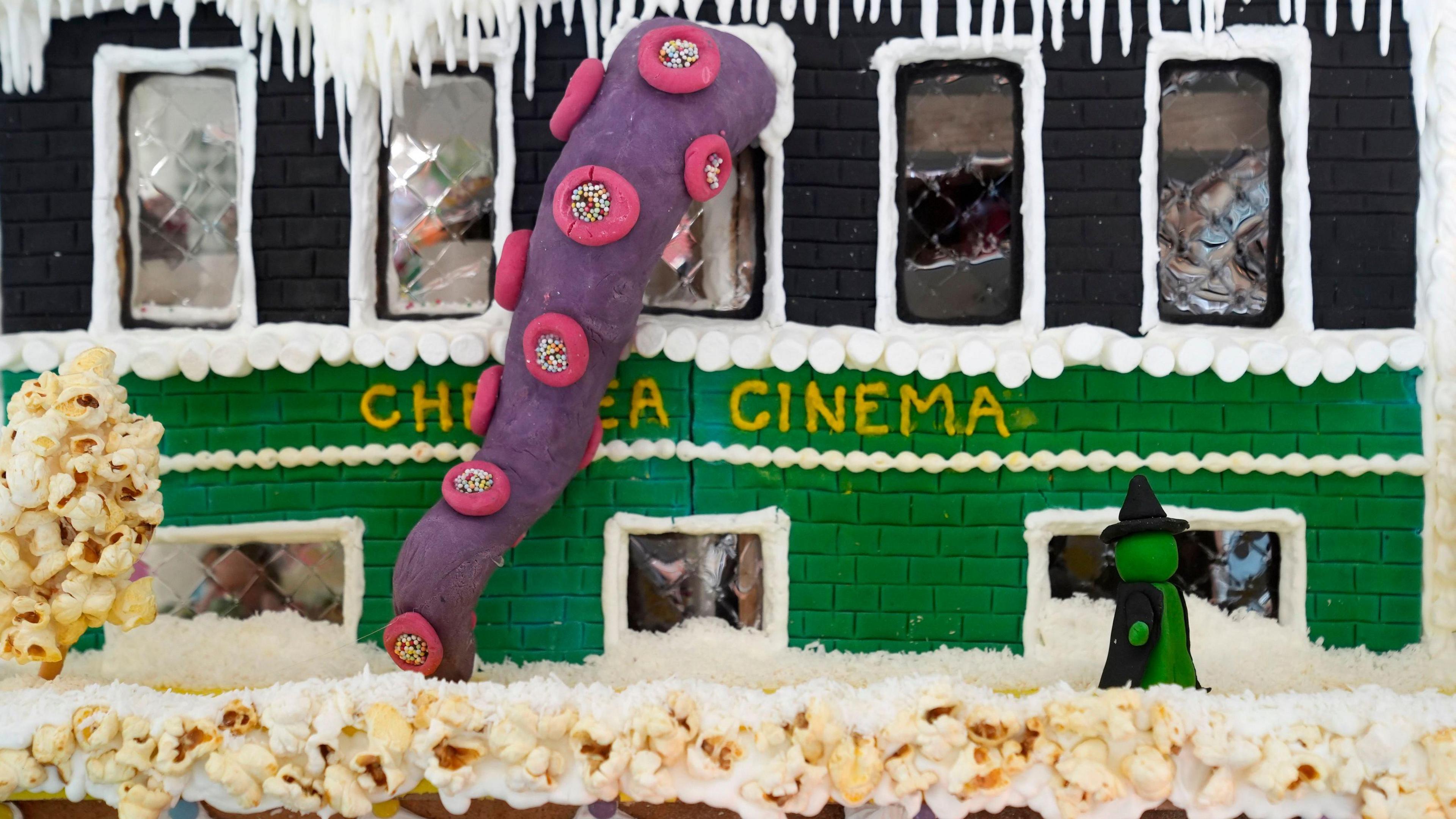 A gingerbread model of The Gaumont Chelsea Cinema. It is green and covered in marzipan, with a large purple tentacle snaking out of a top window. Popcorn has been used to make a layer of snow and a green witch stands on the roof.