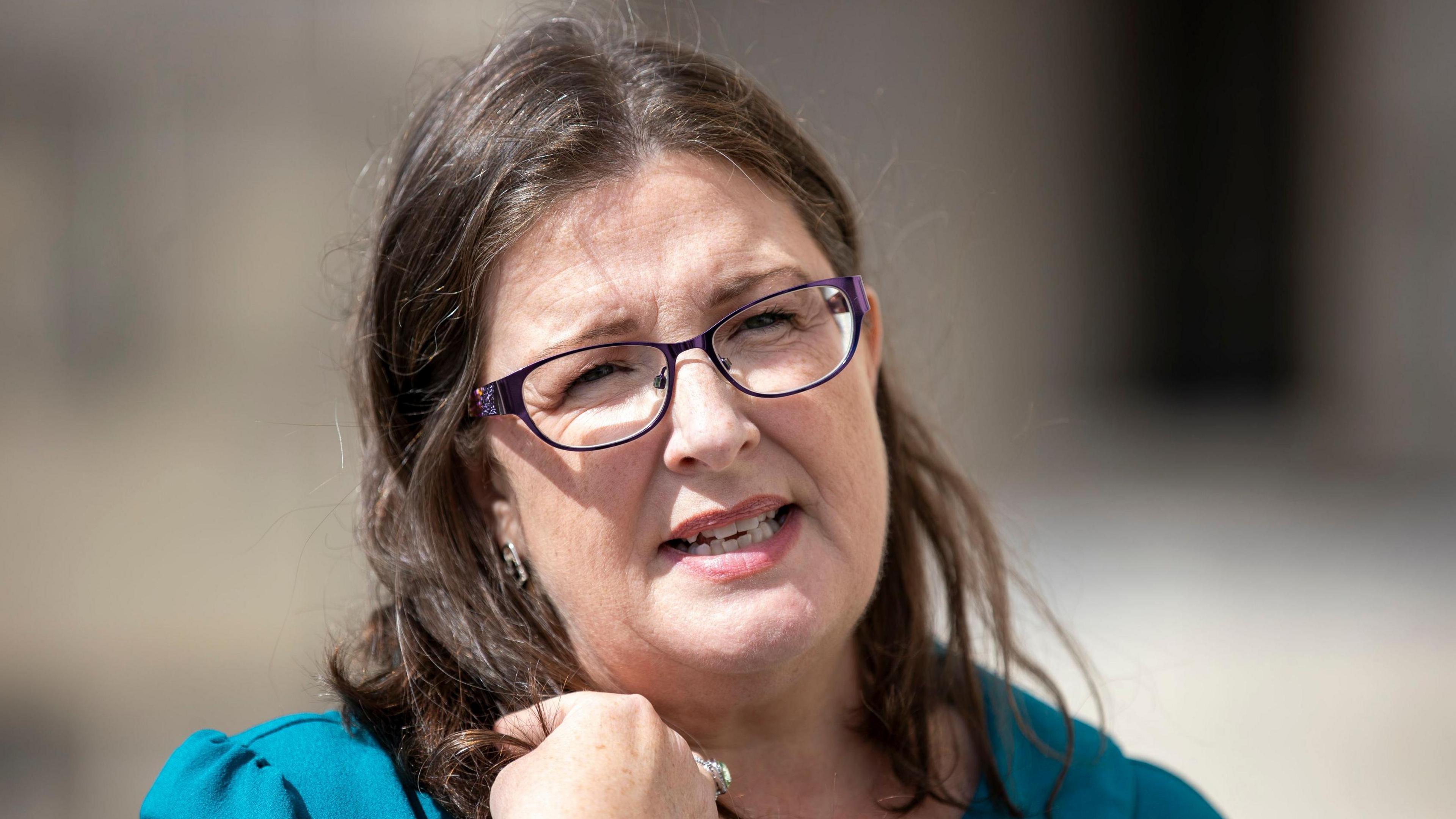 Kellie Armstrong talking to media outside Stormont. She has long brunette hair and is wearing purple framed glasses and wearing a turquoise top. 