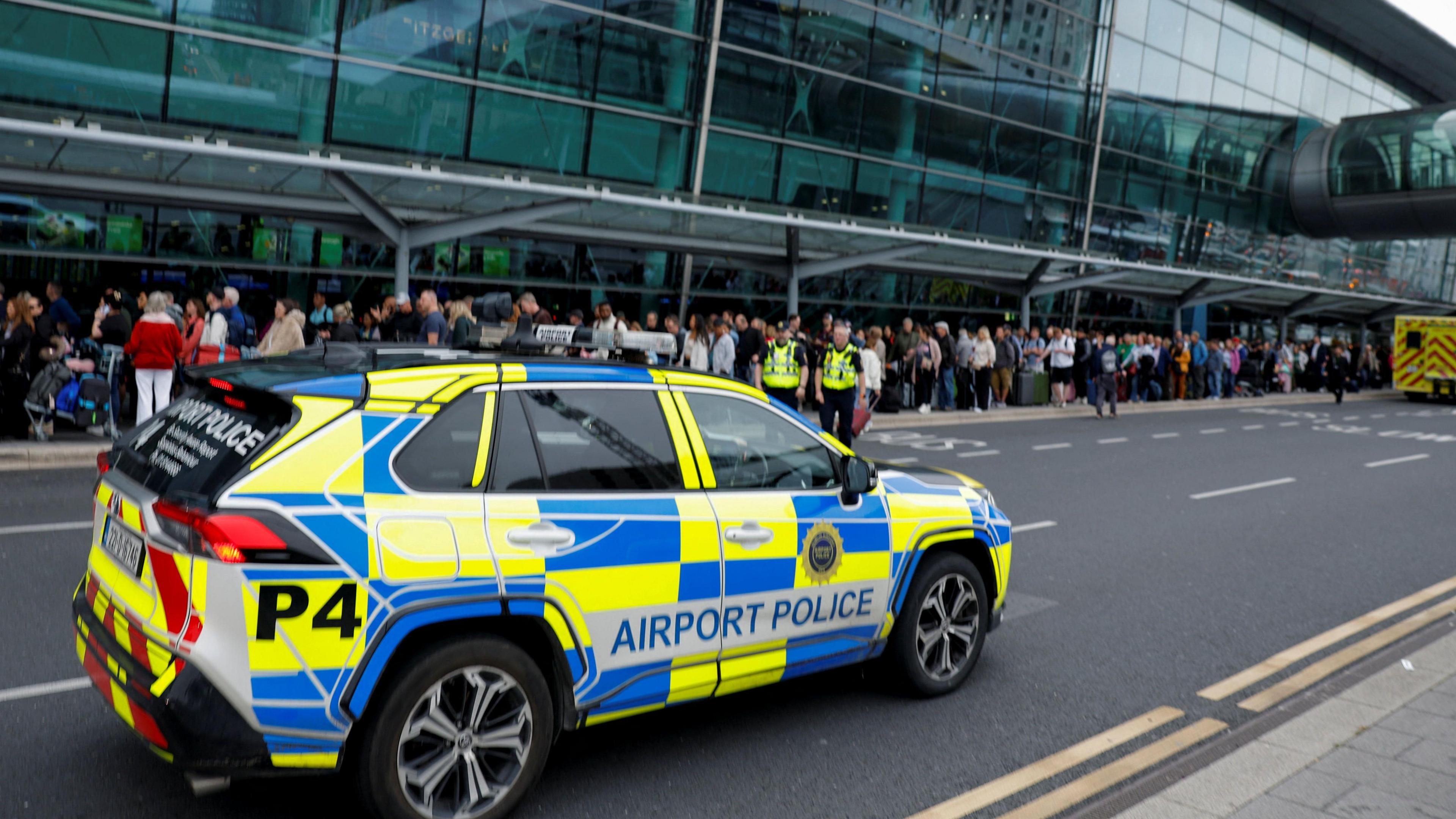 A busy picture, with an airport police car in the foreground. There are two people in high-vis. The back of another emergency vehicle can be seen at the top right. There is also a blurry queue of people lined up outside the glass terminal building.