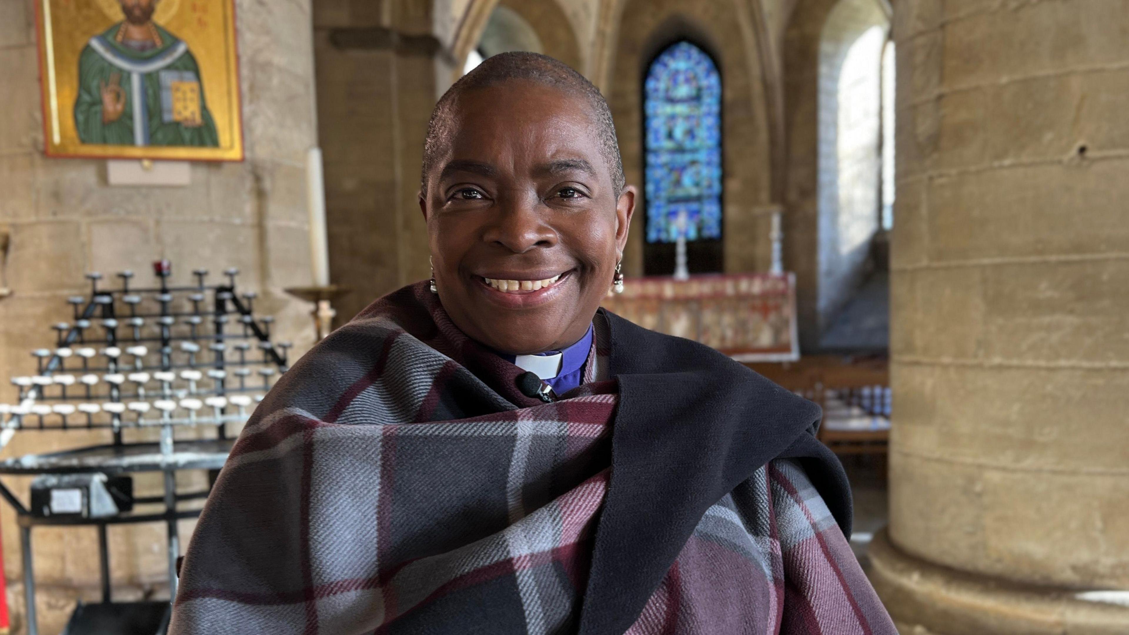 An image of the Bishop of Dover and the Bishop in Canterbury, the Right Reverend Rose Hudson-Wilkin. She is wearing a checked shawl and typical bishop's dress. In the background there is candle display, an image of Jesus Christ and a stained glass window.