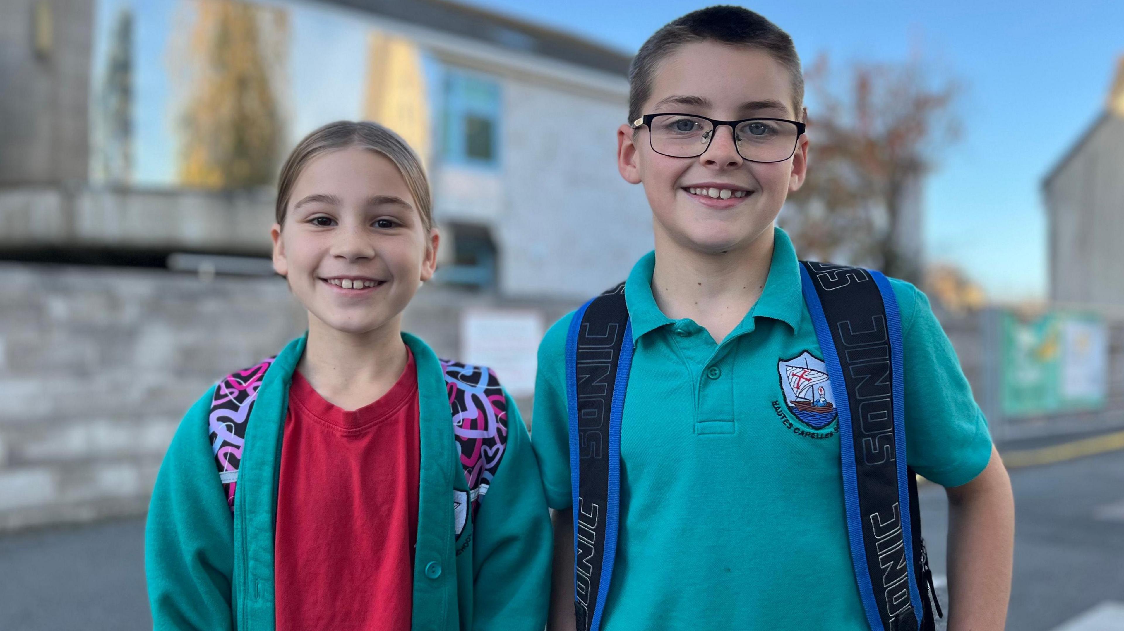 Inara and Sebastian - On the left is a young girl with blonde hair smiling at the camera. She's wearing a turquoise jumper and a red shirt. She has a patterned back back on her back and you can see the straps. On the right is a taller boy with black hair, black glasses and a turquoise polo shirt. He has a backpack on with blue and back straps. 