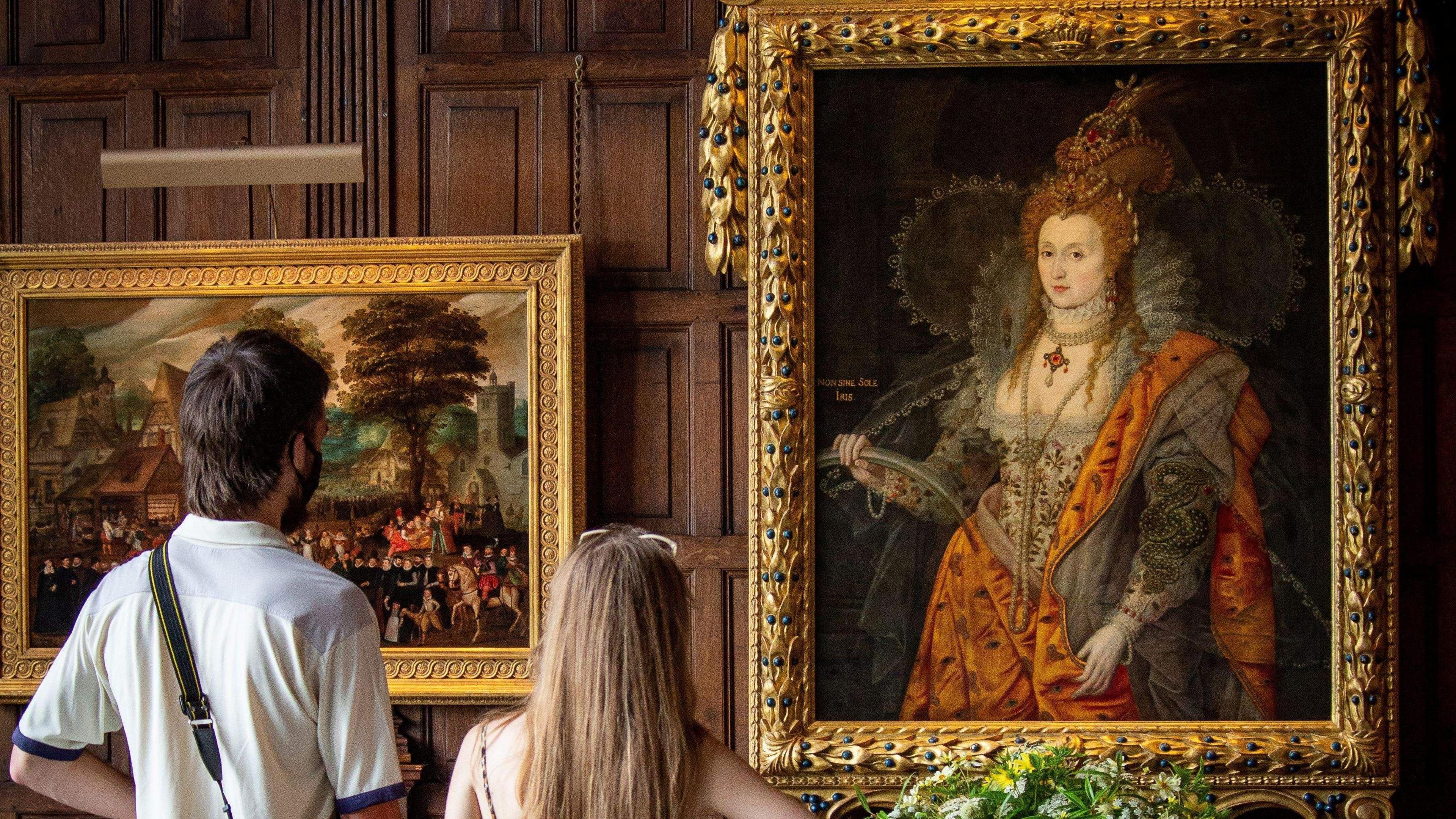 Two people, with their backs to the camera, looking at The Rainbow Portrait, a painting of Queen Elizabeth I at Hatfield House, with another painting next to it