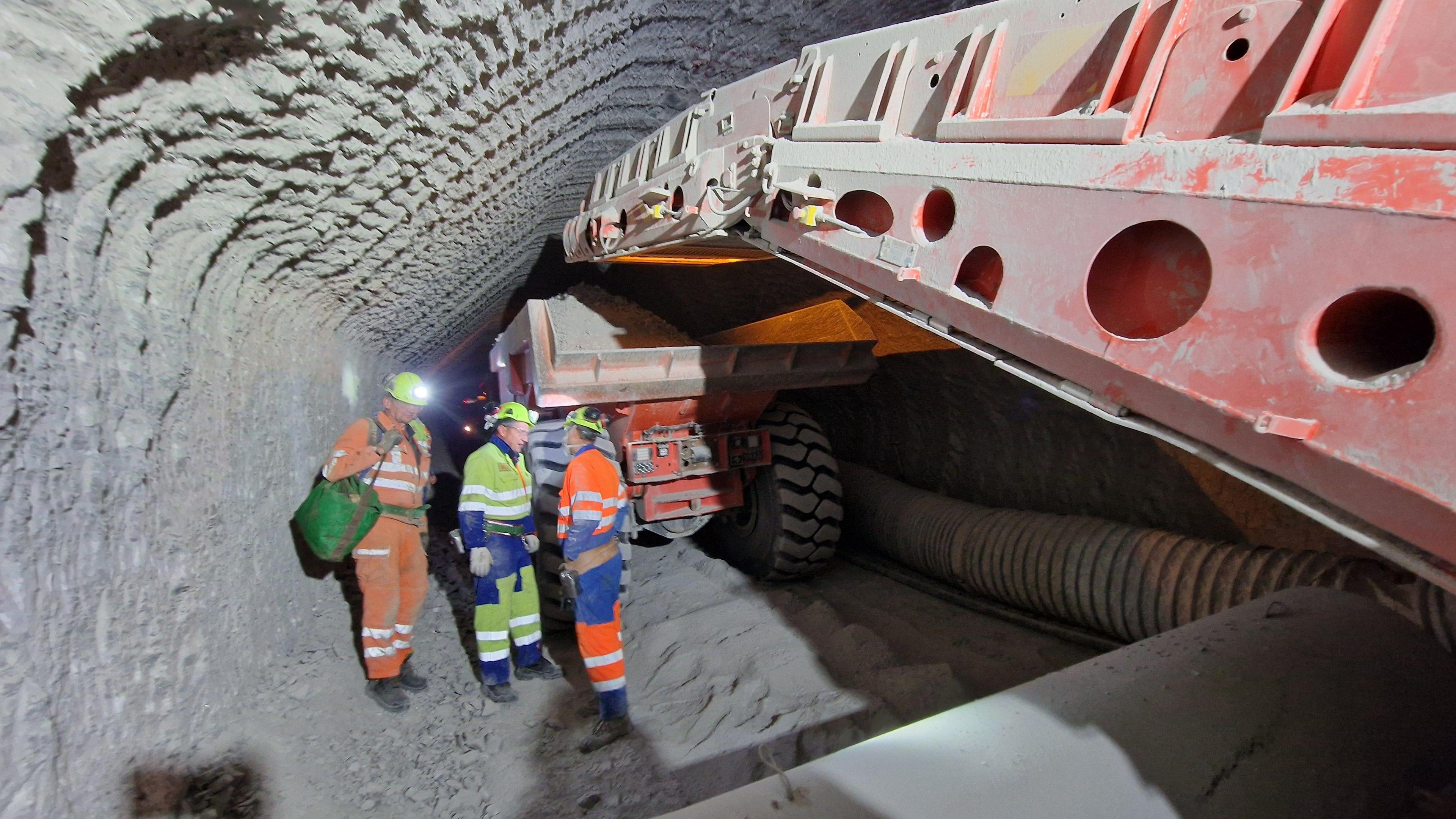 Three men wearing hi-vis jackets stand inside a tunnel in the gypsum mine. There is a conveyor belt in front of a dump truck filled with gypsum.