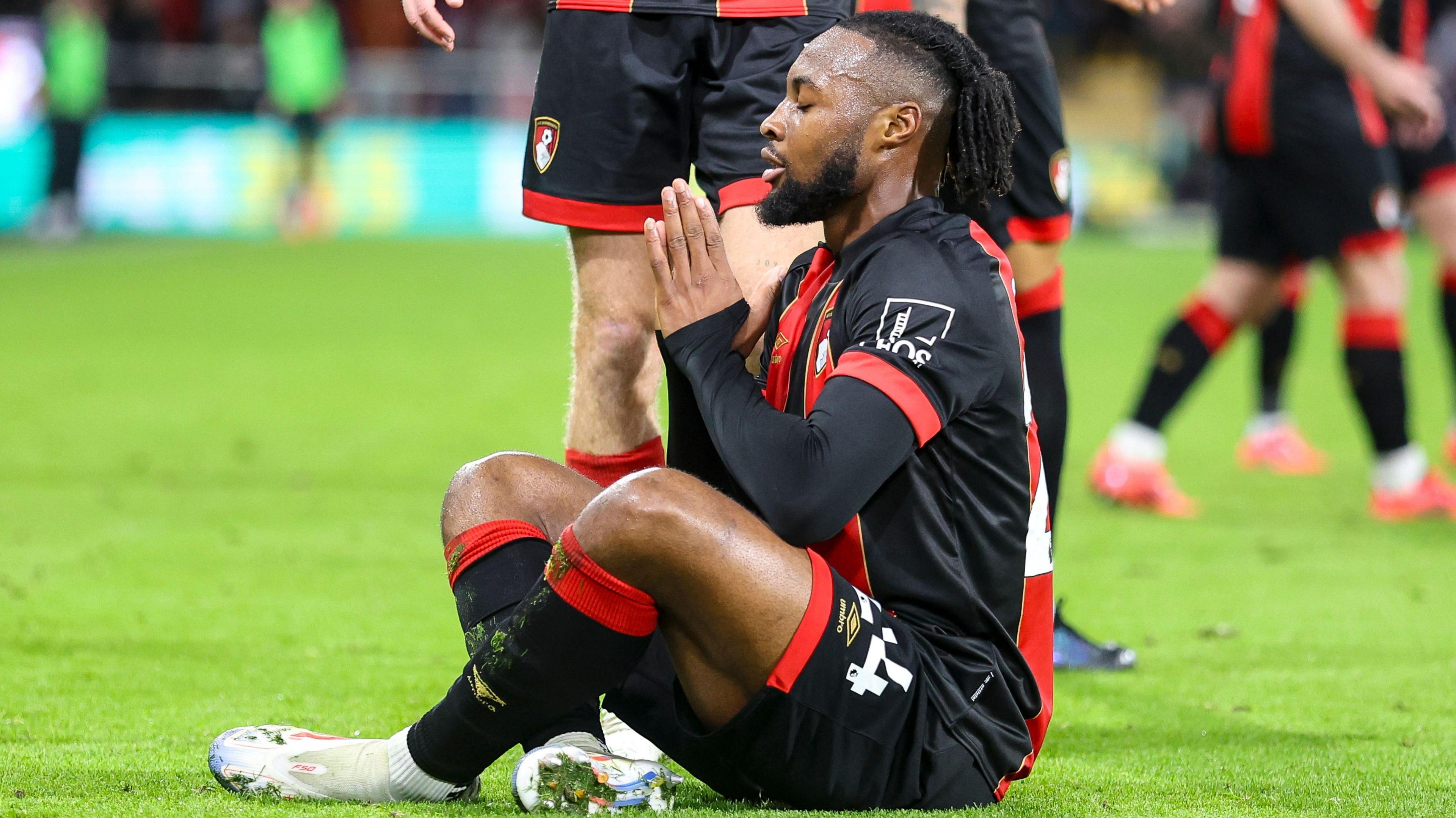 Antoine Semenyo in red and black Bournemouth kit sits cross-legged on the pitch, closes his eyes and brings his hands together in a prayer symbol to celebrate a goal