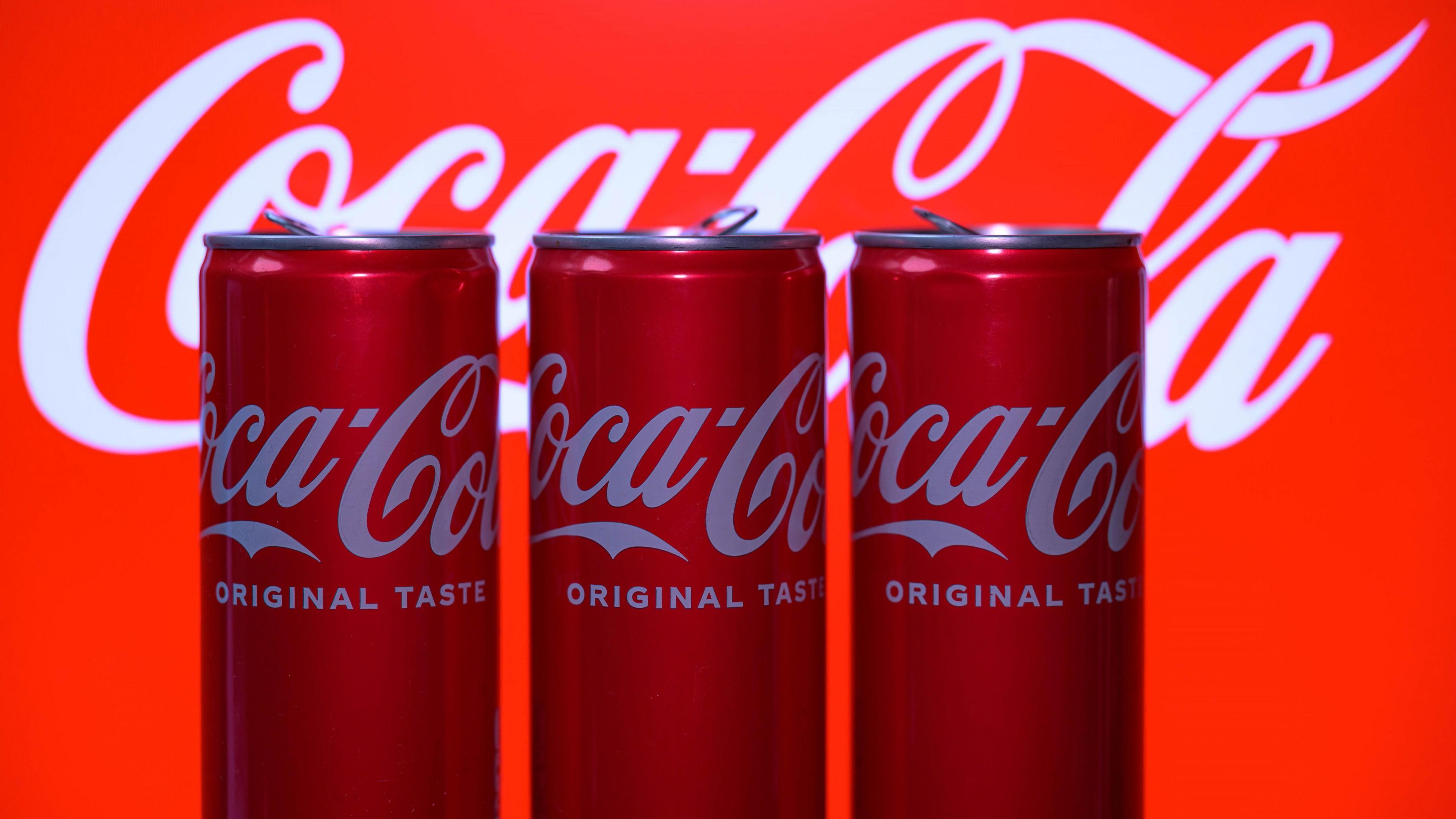 Three Coca Cola cans in a row sitting in front of the large Coca Cola logo  