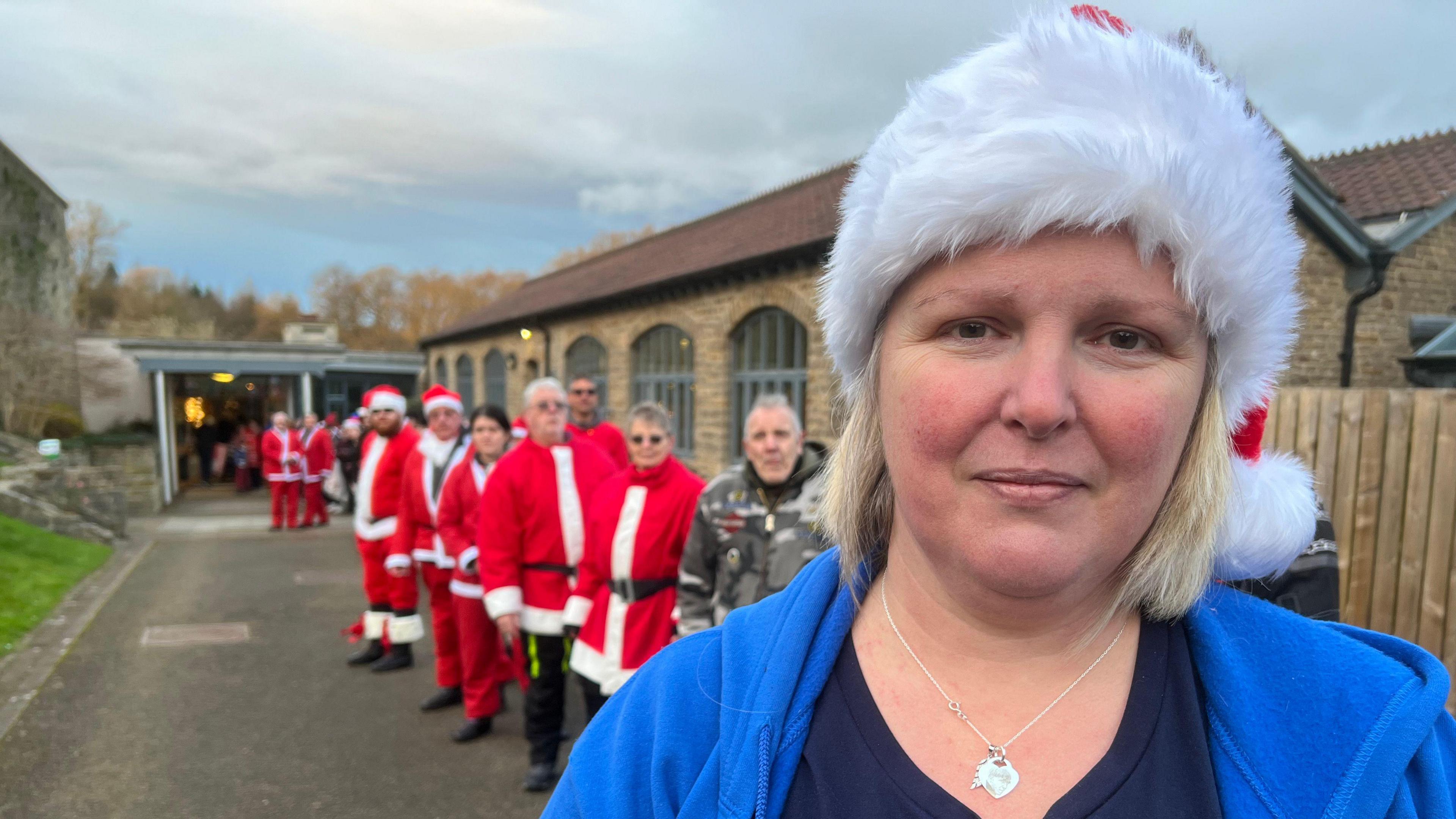 Jenny Gray, wearing a Santa hat and a blue hoodie, standing closed to the camera. A long line of people stand in the background, the majority of them in red Santa costumes. 