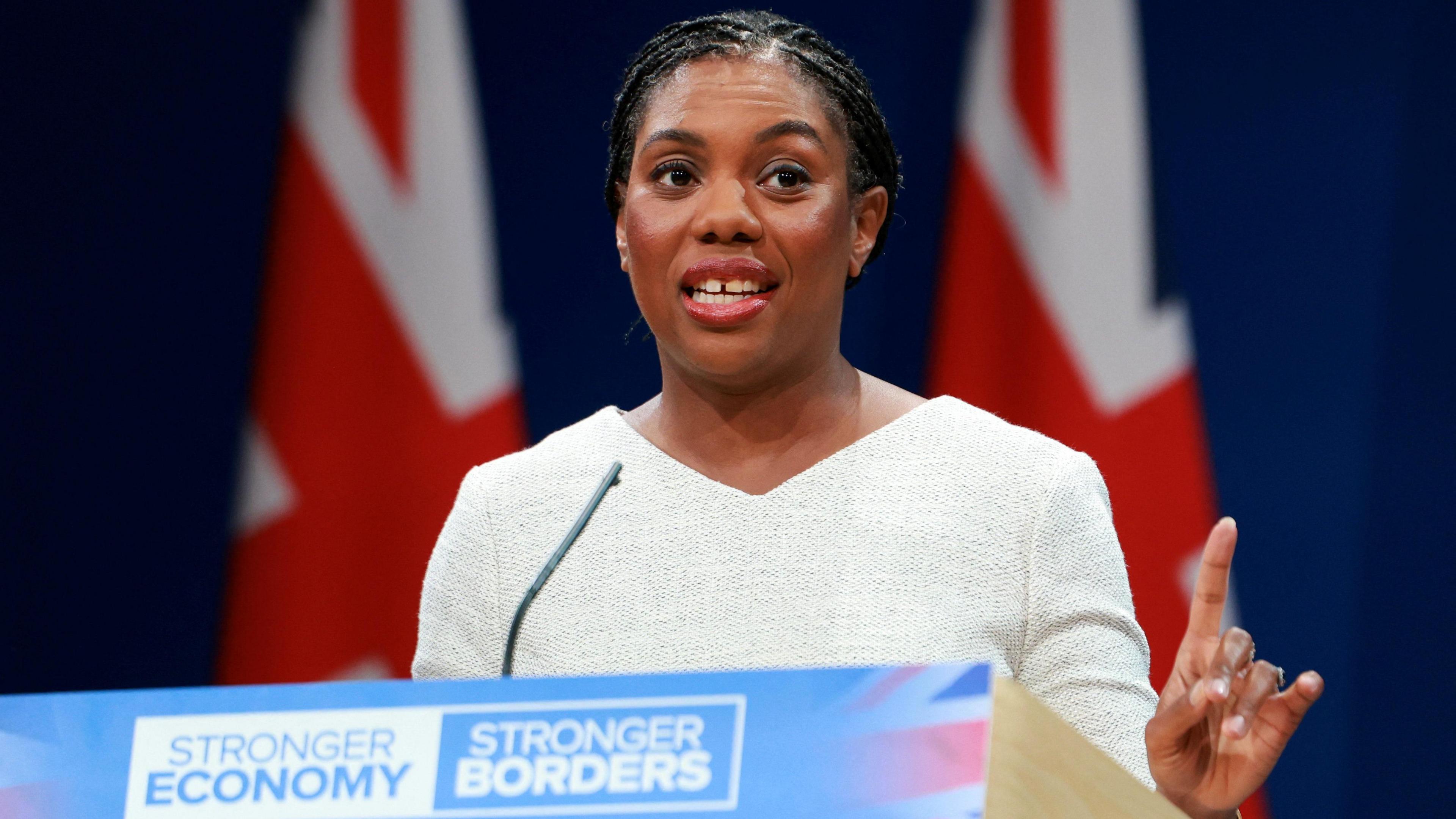 Kemi Badenoch - a woman with black hair tied back and wearing a light grey outfit - speaks at a podium during the Conservative Party conference. The podium is blue coloured with a faded Union Flag on it. It says "stronger economy, stronger border". A microphone is pointing at Ms Badenoch's face as she talks while raising her left hand with the index finger pointing. Two large Union Flags are displayed behind her.