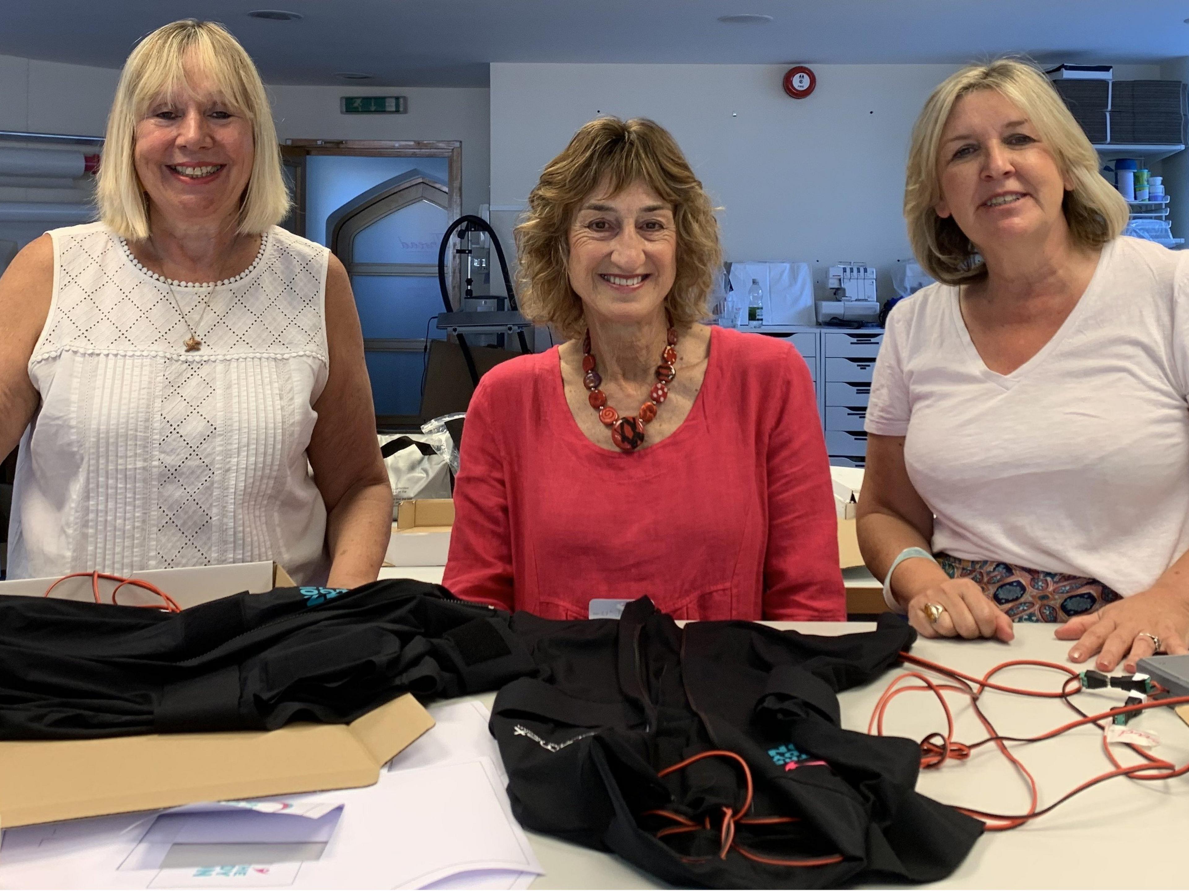 Three women stand in front of a desk. On the desk is a black body-warmer style vest, with electric heating pads inside.