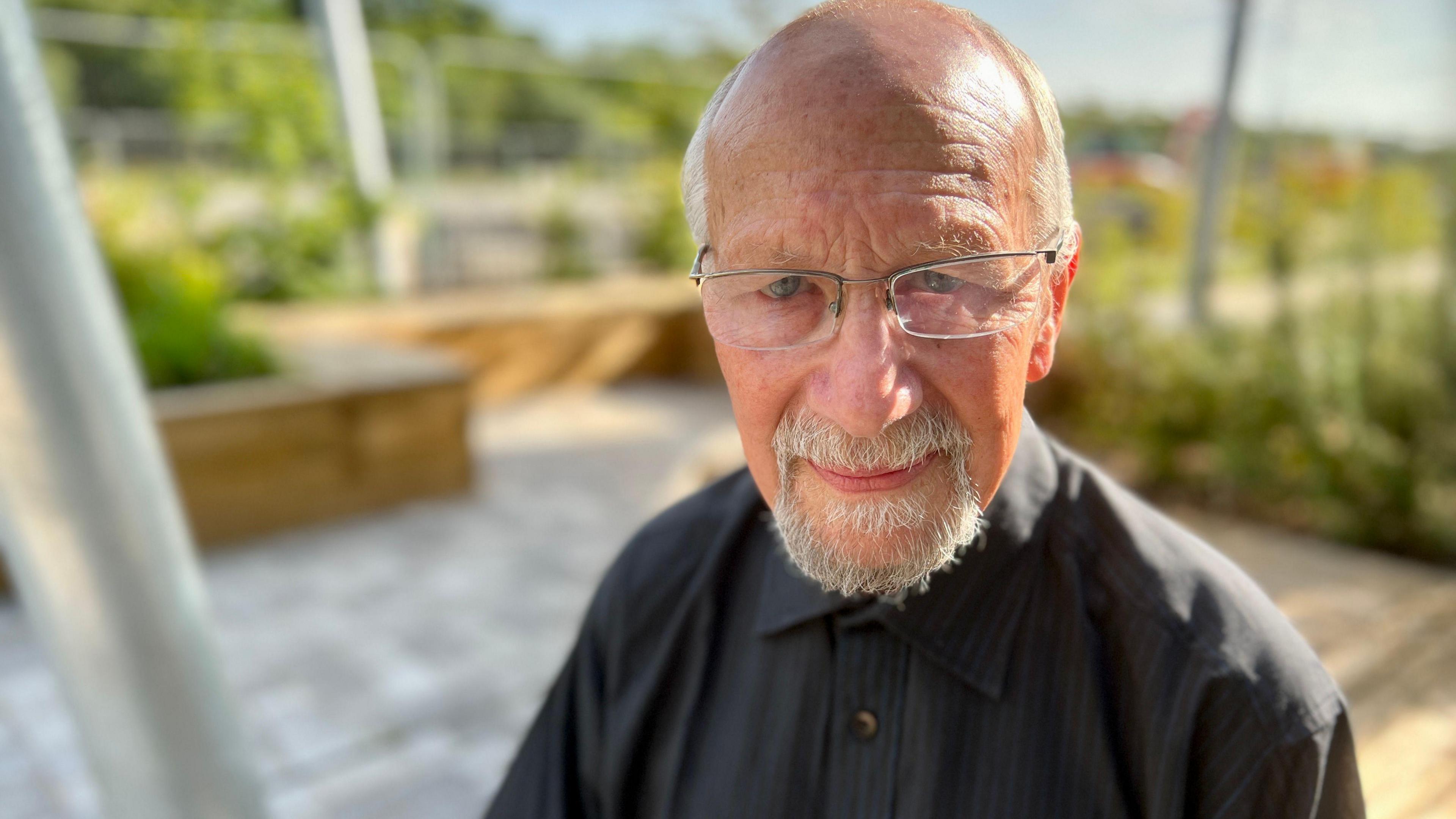 John Newman looks at the camera. His head and shoulders are visible. He is wearing a black shirt and has glasses on. He has blue eyes and a tanned complexion with a bald head and some grey hair to the side with a grey moustache and beard.