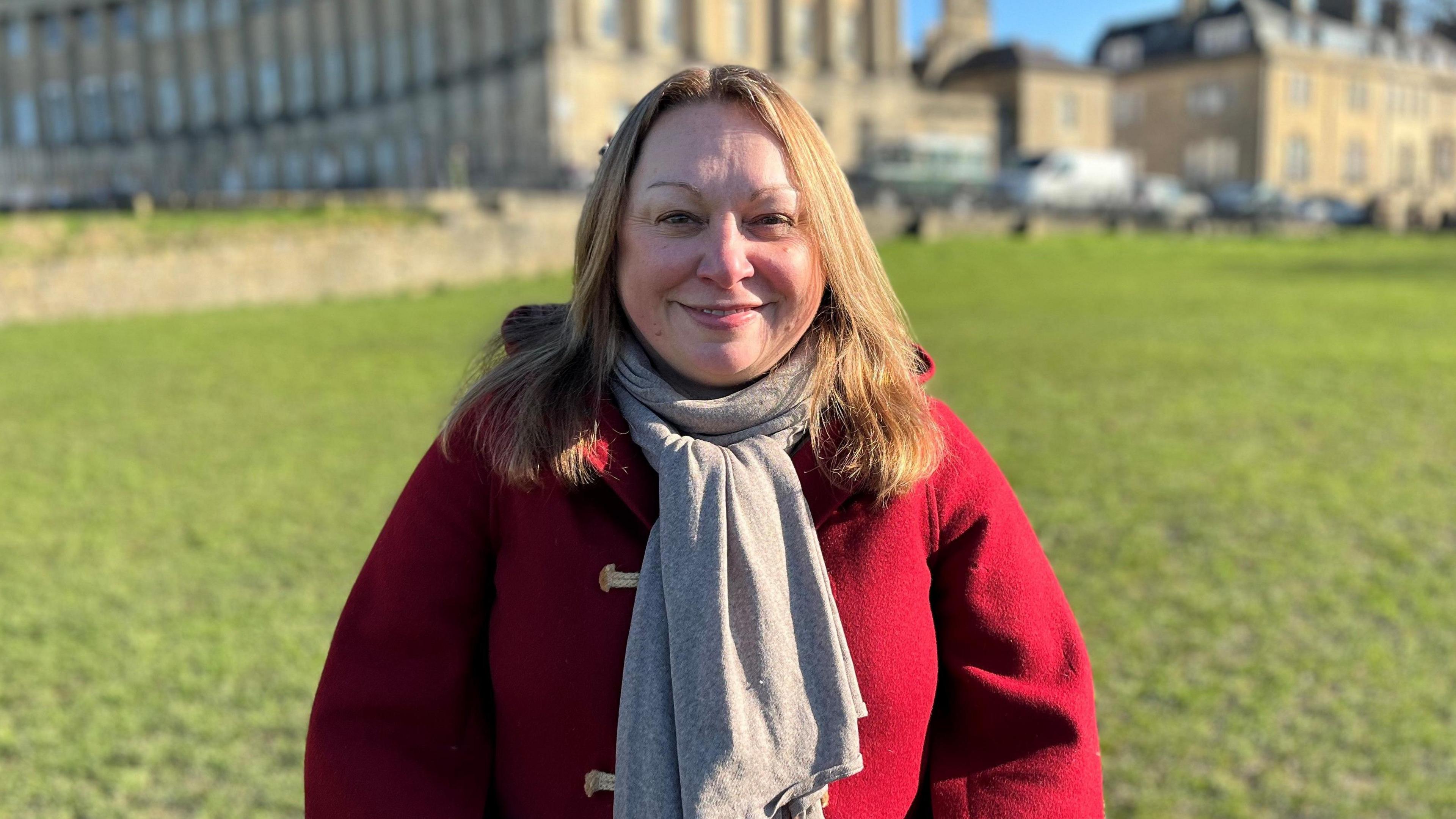 Penny Agent wearing a red coat with wooden toggles and a grey scarf. She has medium length strawberry blonde hair and is smiling at the camera. It is a bright sunny day, and she is standing on the grassy field in front of the Bath Crescent, which you can see in the background out of focus.
