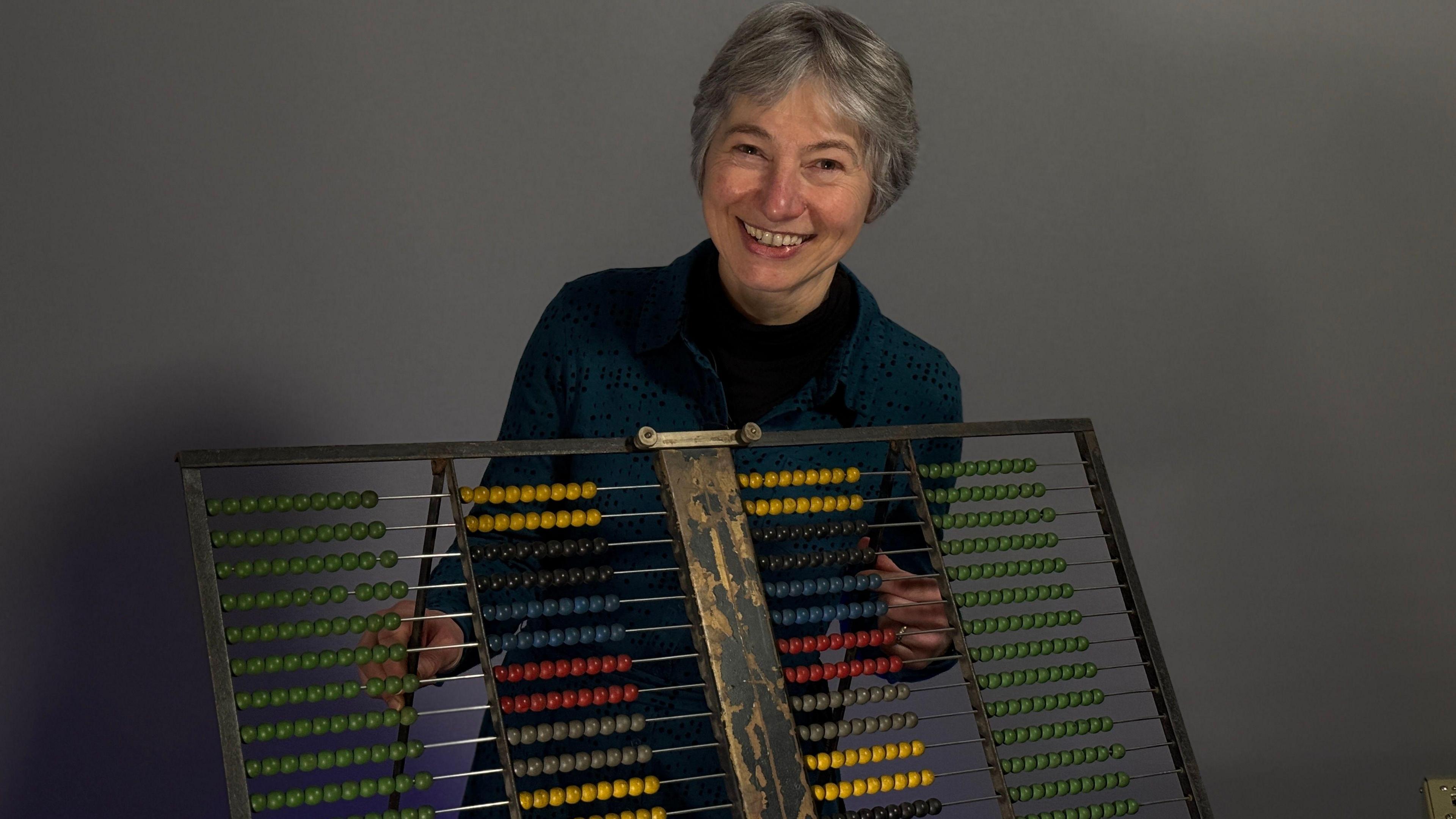 Gillian Cooke stands behind an large abacus, about 80cm wide. It has many rows of different coloured beads which were used by examiners to set the grade standards.