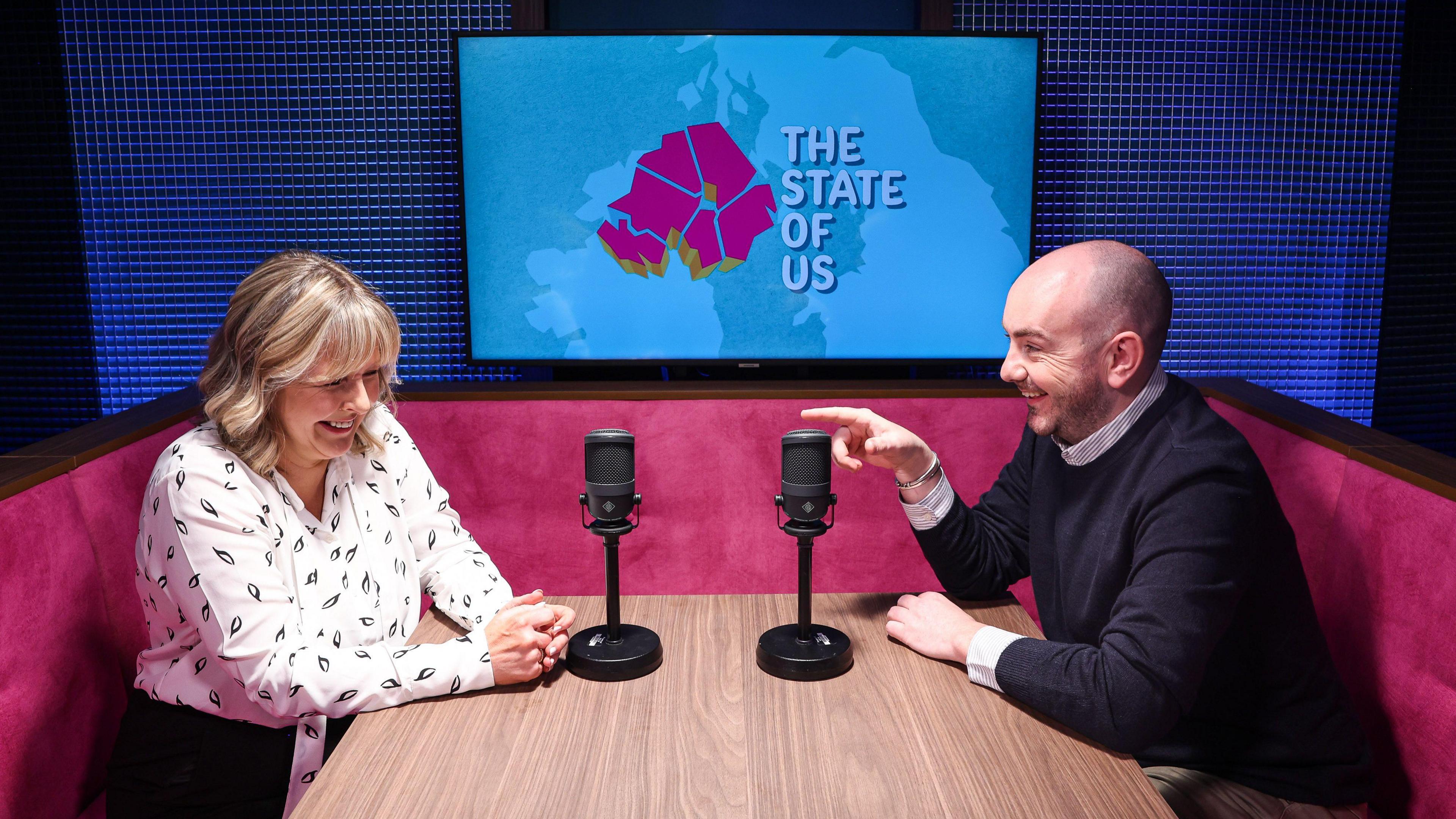 Declan Harvey points towards Tara Mills who are both sitting in a podcast Studio. There is a television screen with the special logo for the state of us podcast visible on a wall in the background. They are seated on a pink sofa. Tara is wearing a white blouse and Declan has a black jumper on.