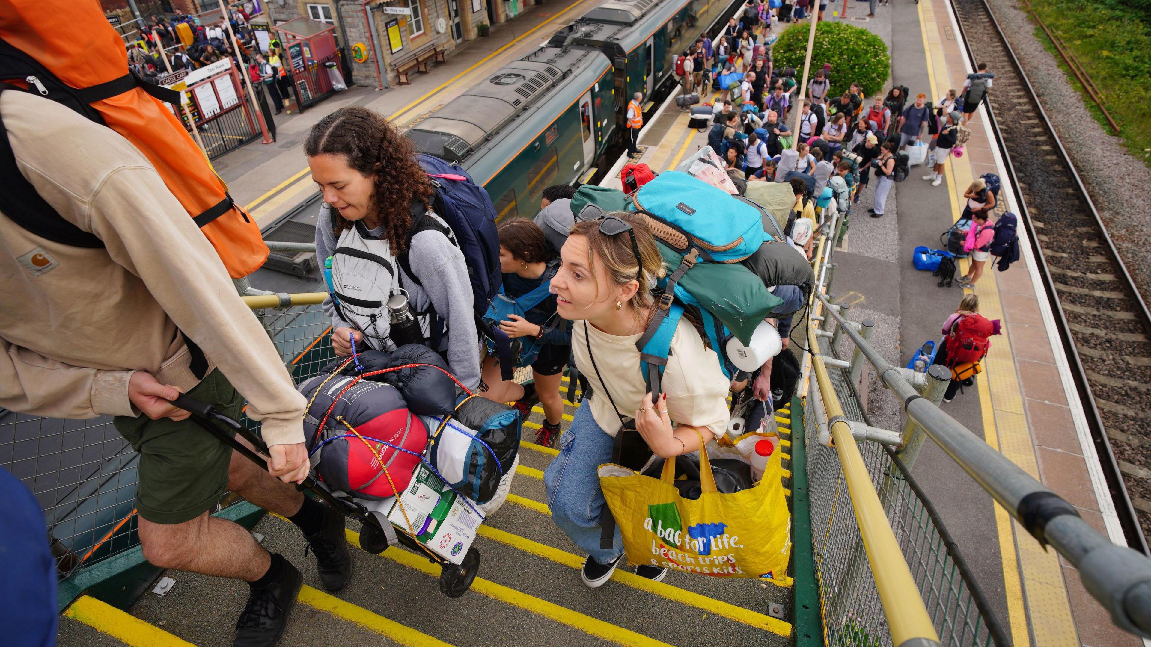 A large group of people disembark a train and walk up a railway bridge carrying large bags.
