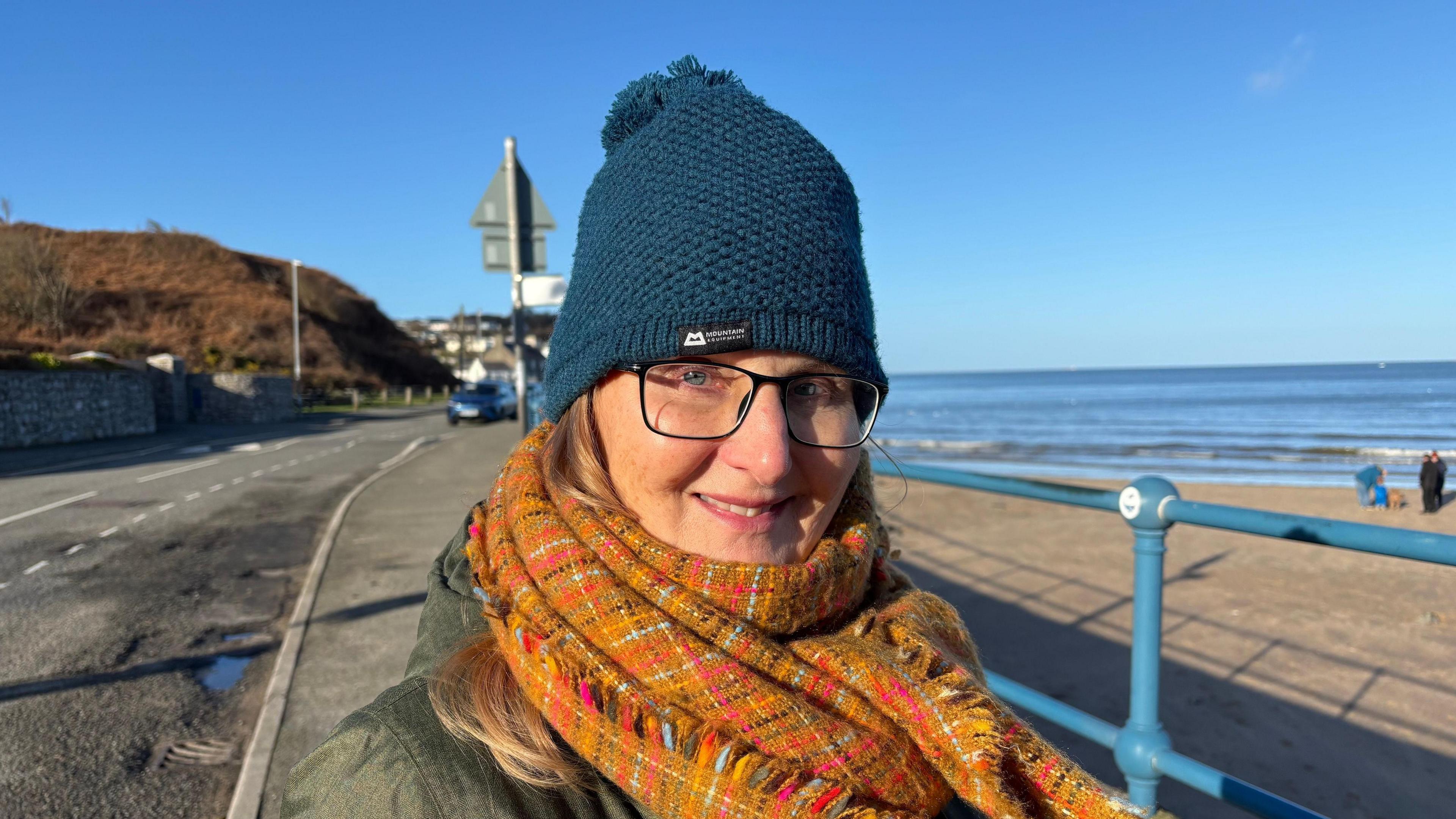 A woman is wearing a dark blue woollen hat with a blue pom-pom just visible, with a orange and yellow plaid checked woollen scarf with blonde hair peeking out from her hat. She is wearing glasses and a green top. 