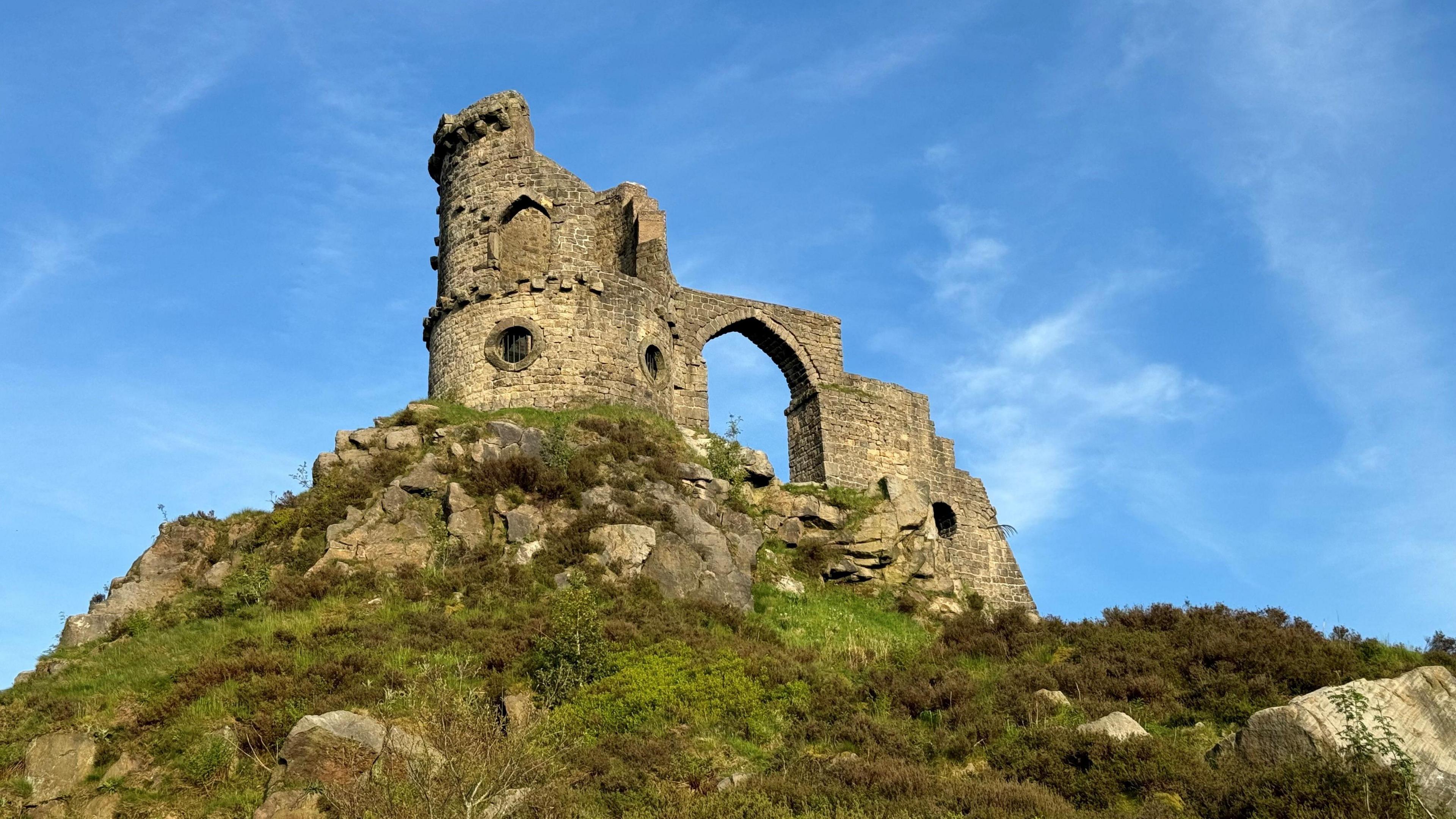 A brick-built structure resembling a derelict castle is stood on top of a hill, with blue sky behind and lush undergrowth beneath it.