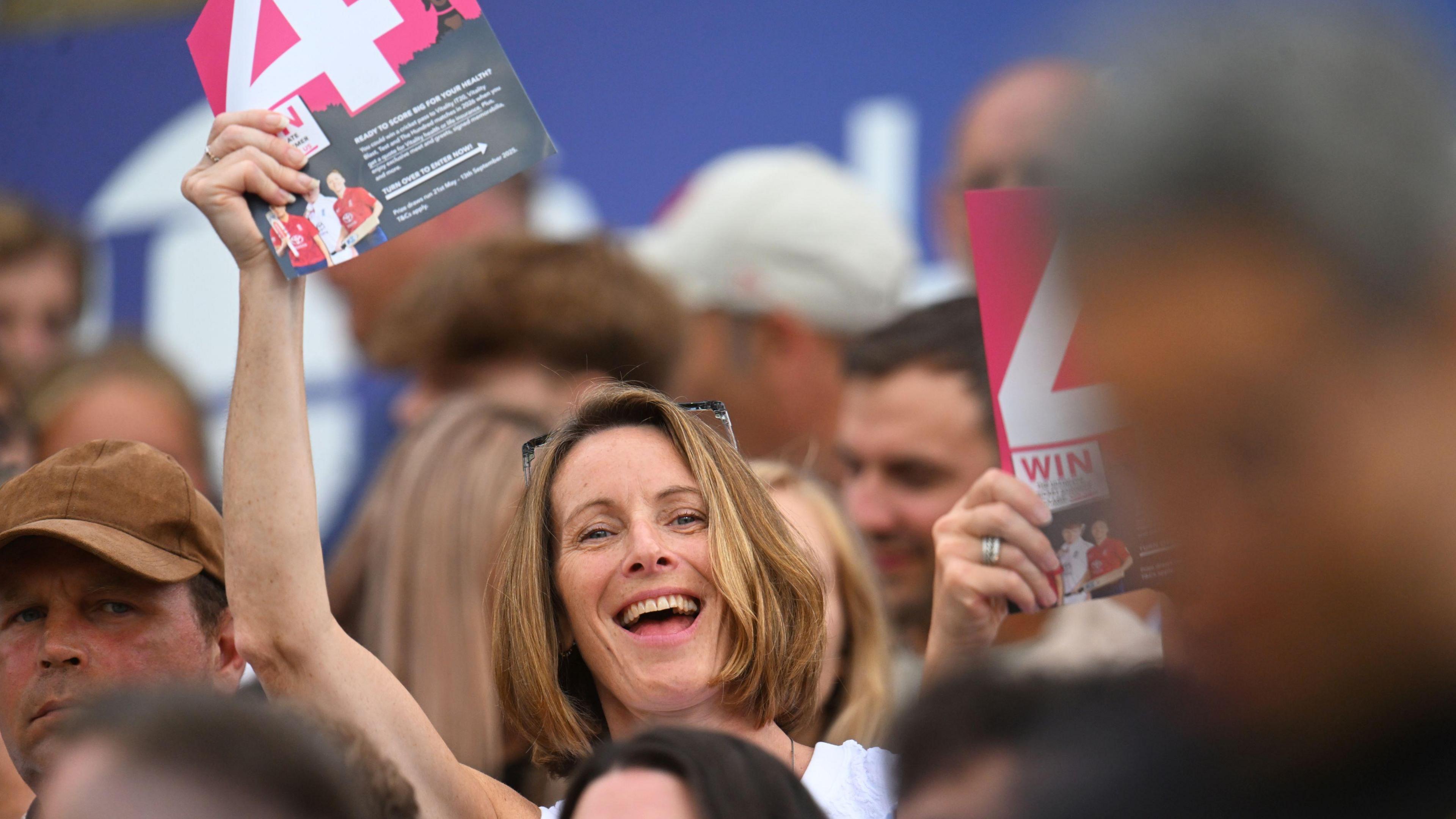 A woman holds up a placard with the letter 4 on it as she watches Somerset v Gloucestershire in the T20 Vitality Blast at Taunton. She is smiling at the camera and has a shoulder-length bob of brown hair. Around her are other fans