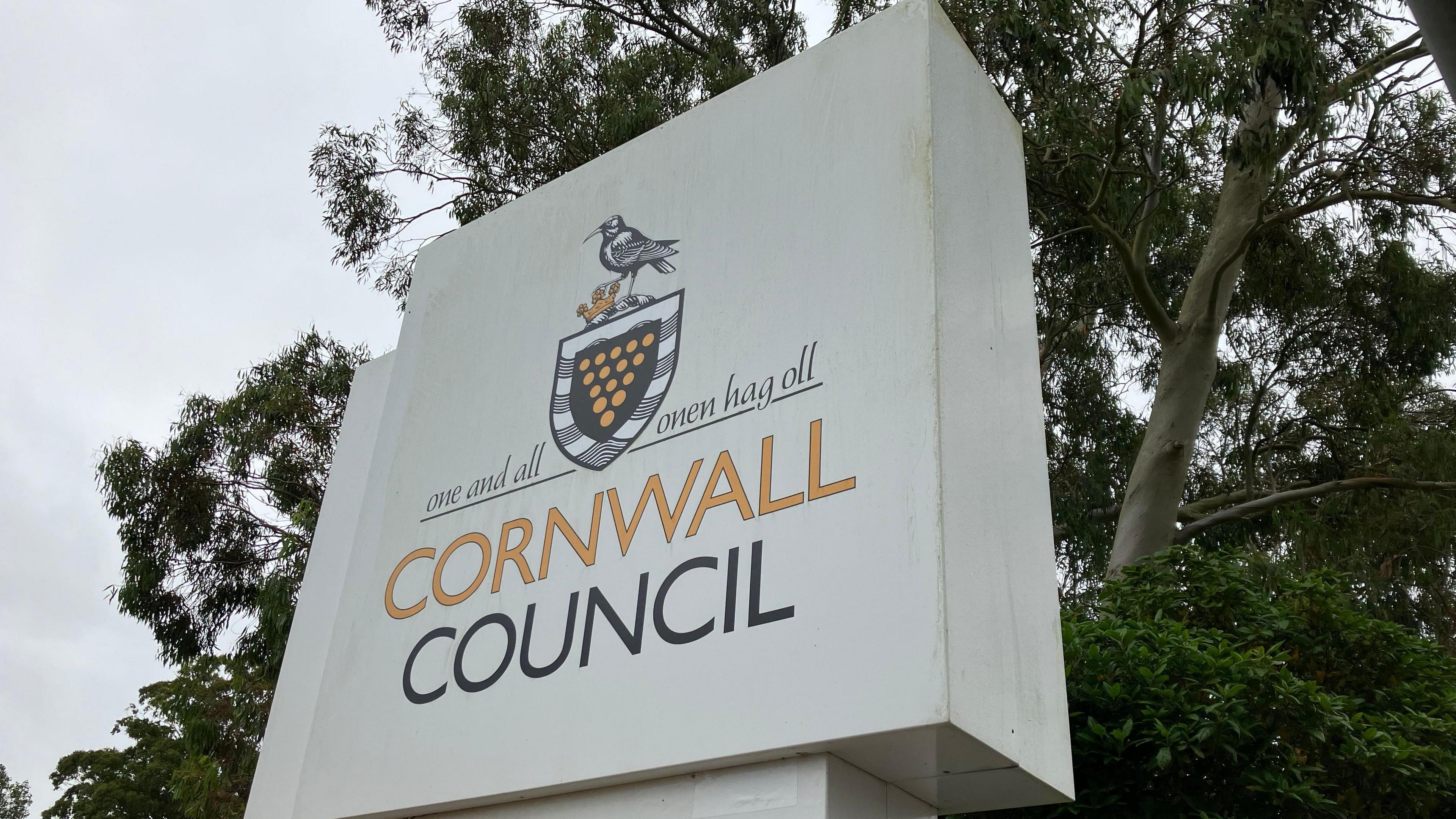 A large white sign with the words 'Cornwall Council'. There is a badge above the text. Behind the sign are trees.