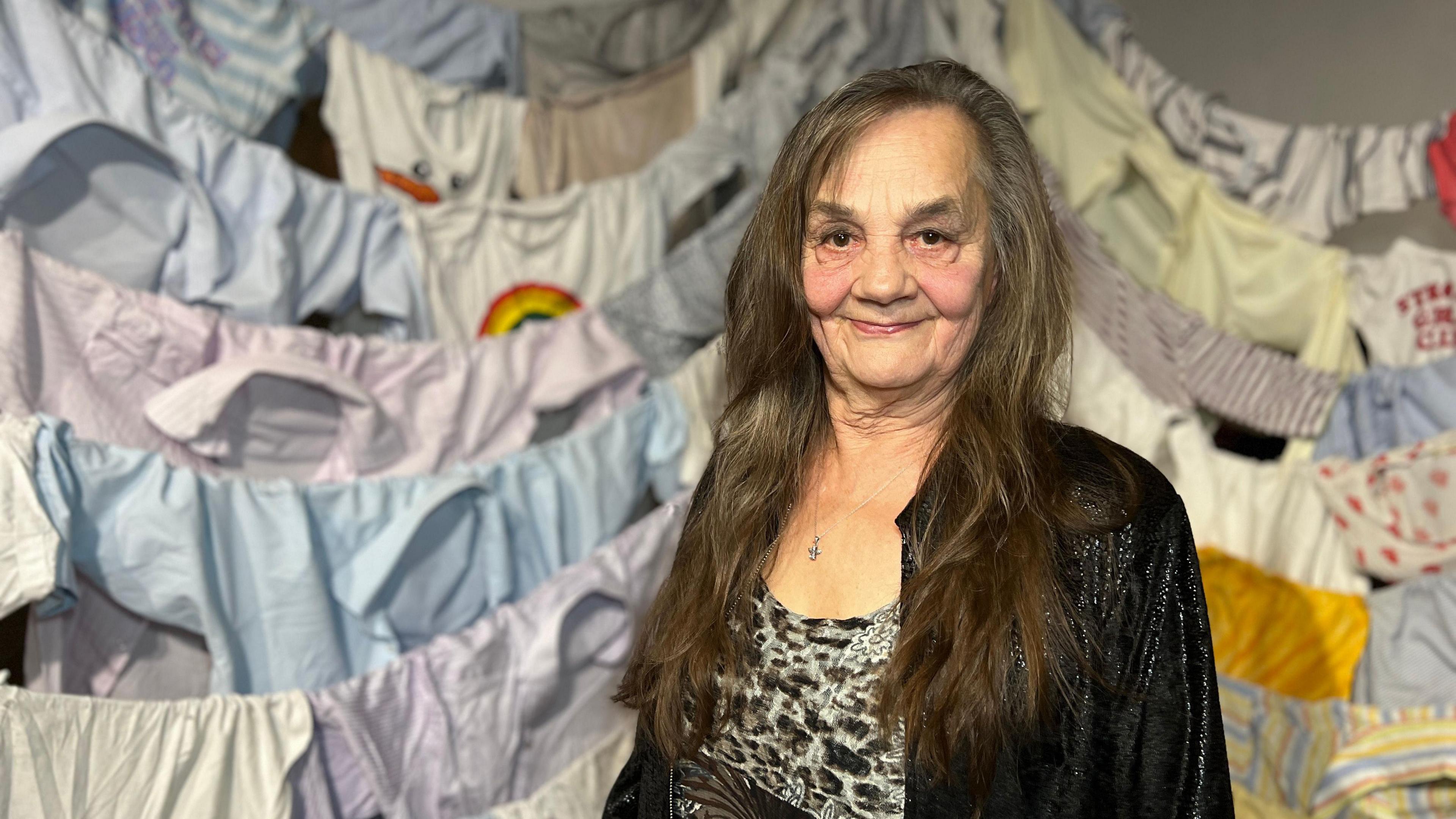 Kaarin looks at the camera as behind her, hundreds of shirts are hung on lines. Kaarin is wearing a black jacket over a leopard print dress and she has long dark hair.