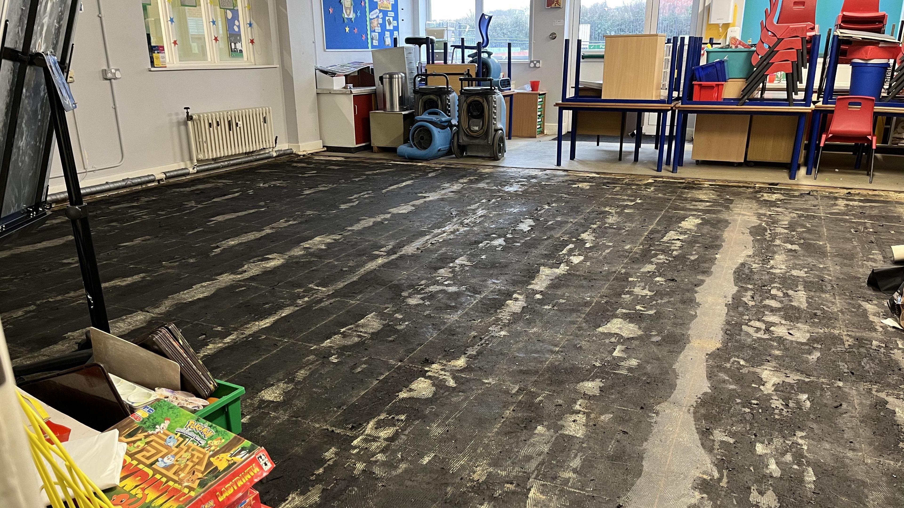 A bare floor in a classroom with all of the furniture moved to one side of the room.