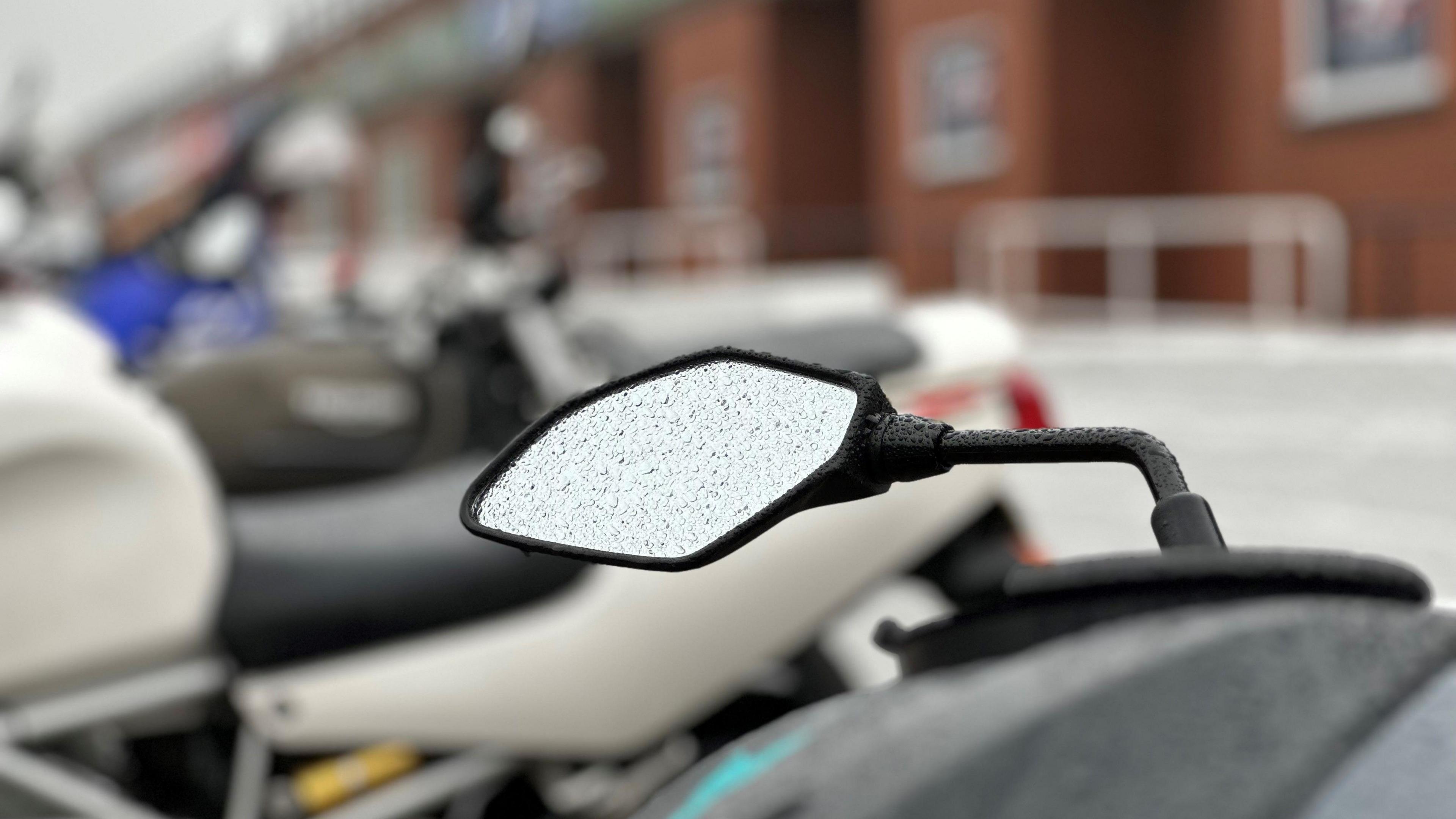 The mirror of a motorcycle with rain drops on it. It's in a row of race fans' motorcycles parked in front of the grandstand.