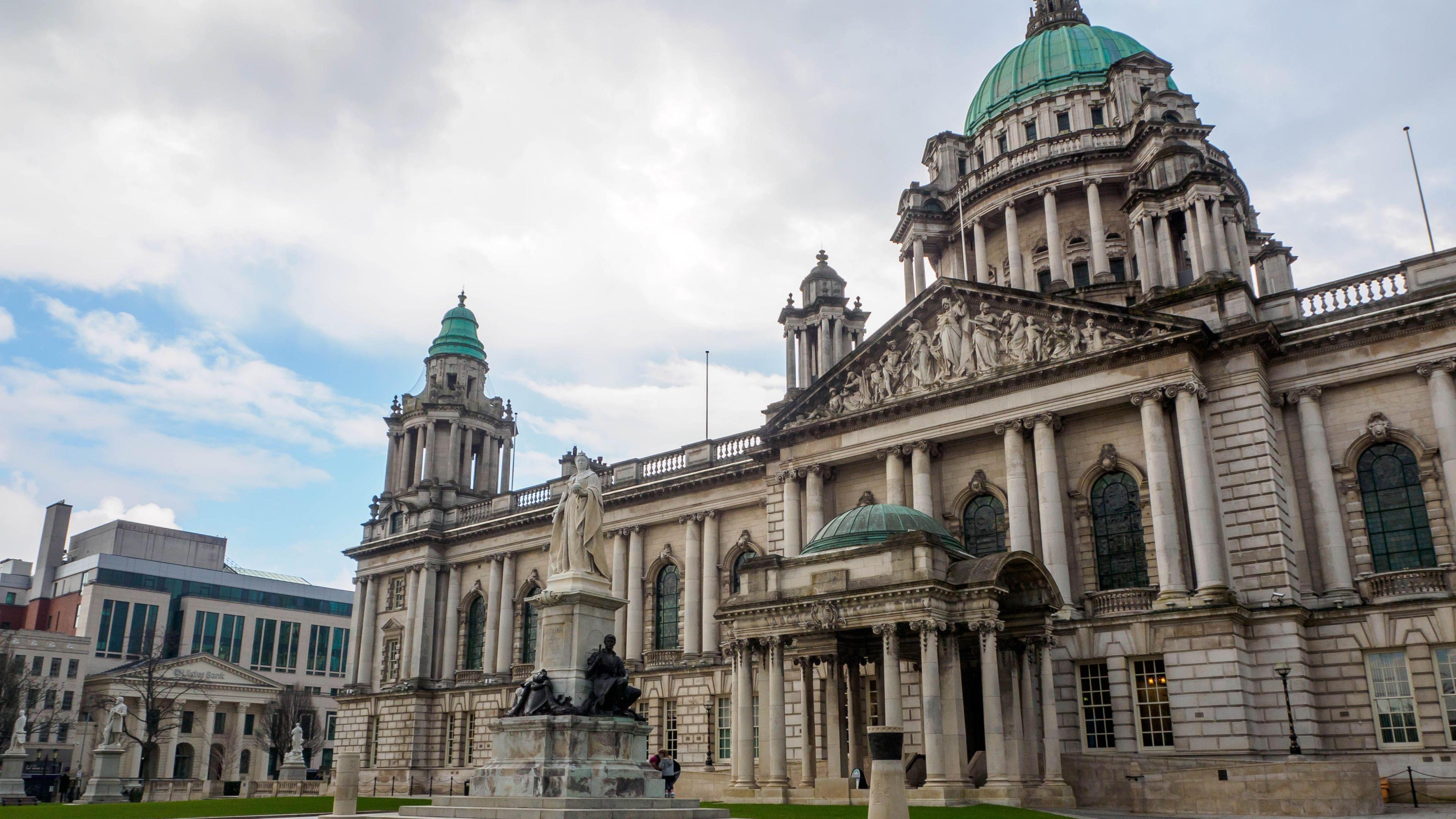An exterior shot of Belfast City Hall. It is a large, imposing building made from blocks of light coloured stone and has a green dome on its roof. There are also a number of statues on the lawn in front of the building.