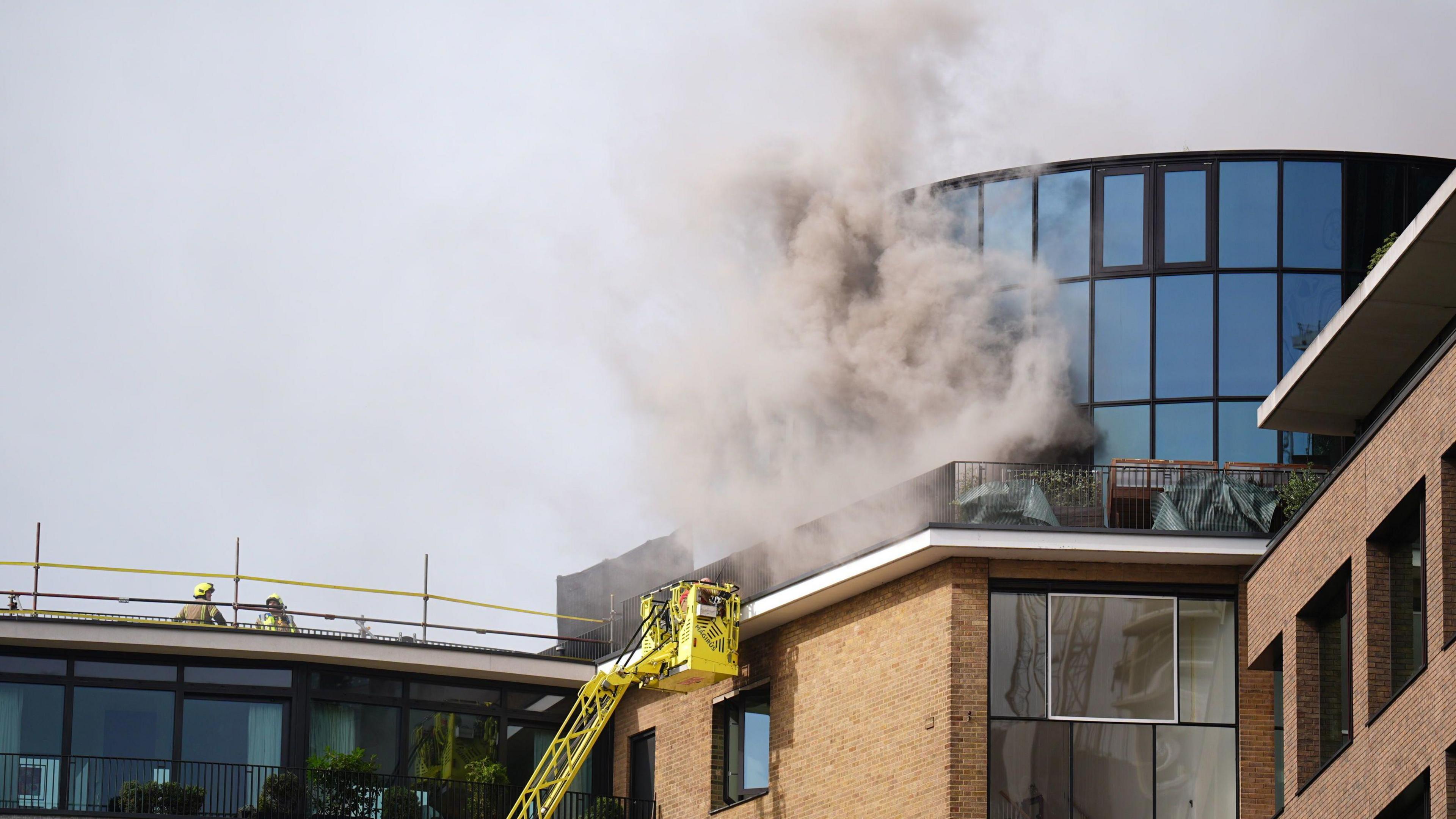 A plume of smoke comes out of a building, with firefighters on scene. 