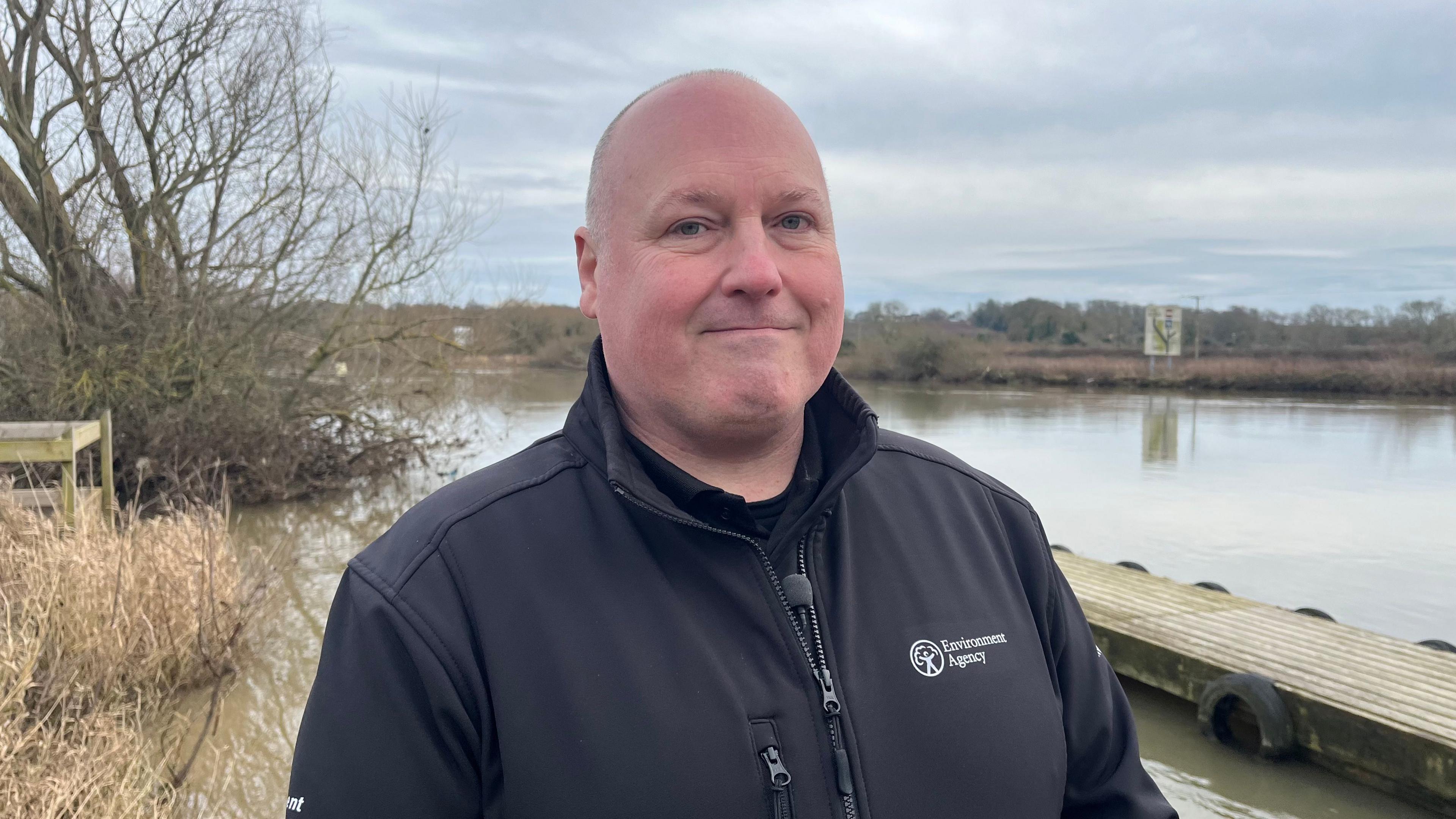 Matthew Gable from the Environment Agency stood next to a river. He has a bold head and is wearing a black jacket with an Environment Agency logo on.  